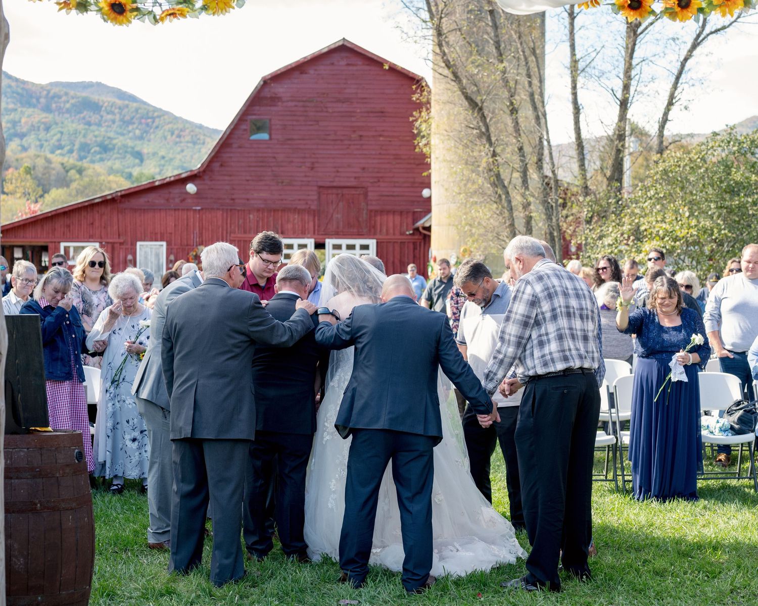 Wedding ceremony blessing outdoors near red barn with mountain backdrop and sunflower decorations.