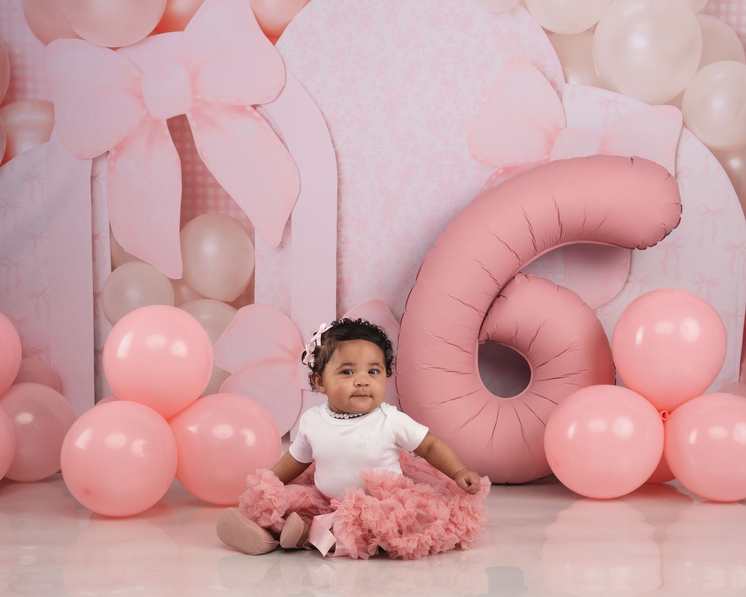 A baby girl in a pink tutu sits before a pink balloon and gift box backdrop with a number six balloon.