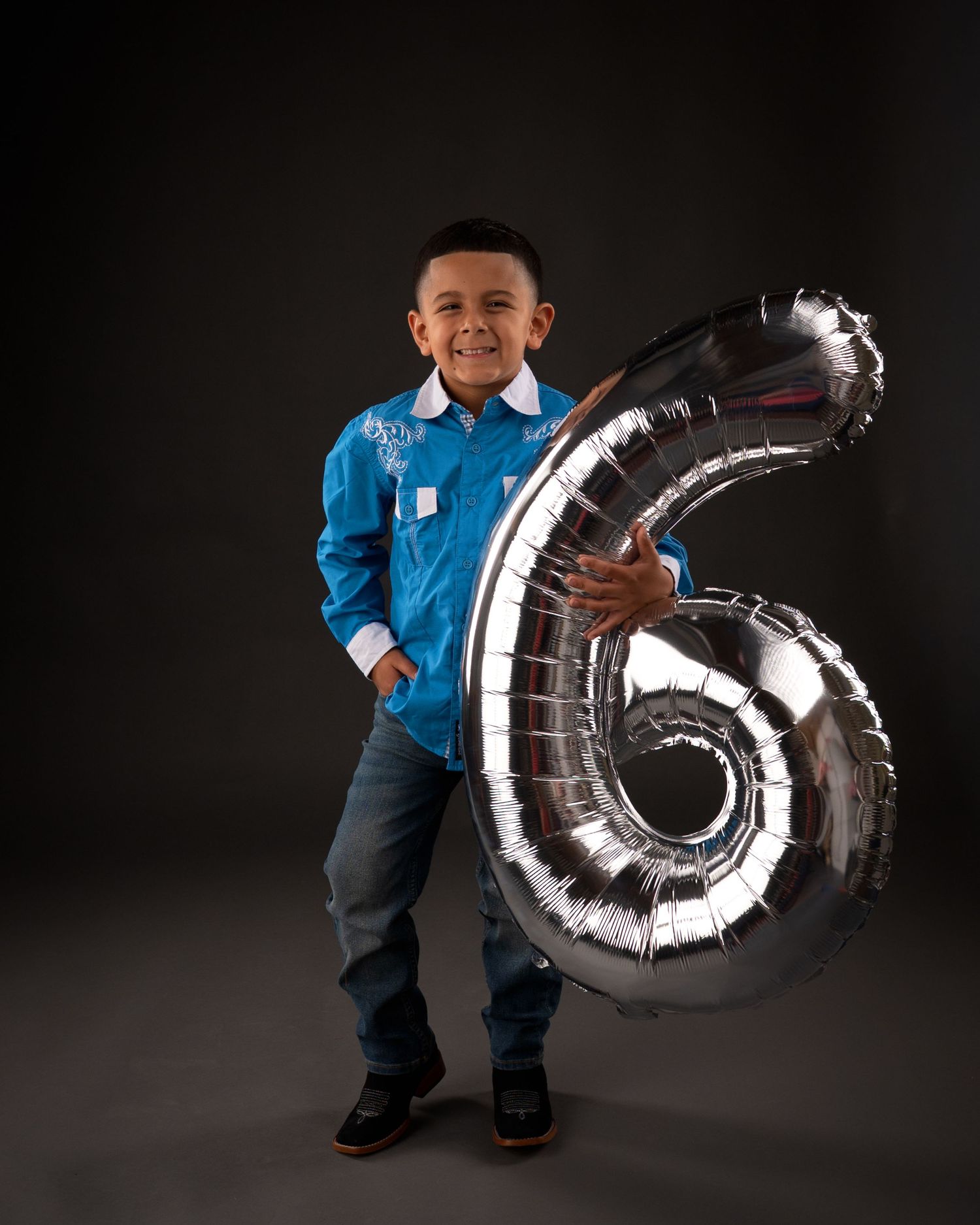Smiling boy in blue shirt holds large silver number 6 balloon for birthday portrait.