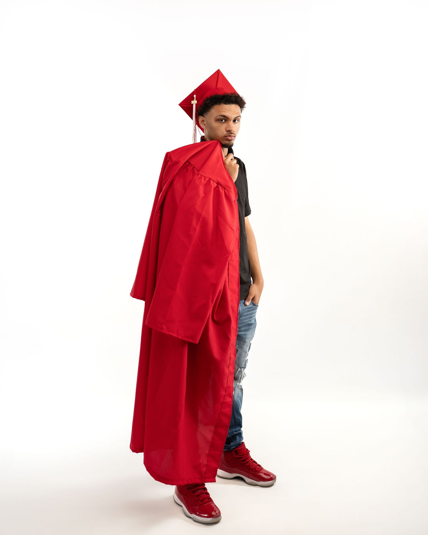 Male graduate in red cap and gown poses sideways in studio with red sneakers.