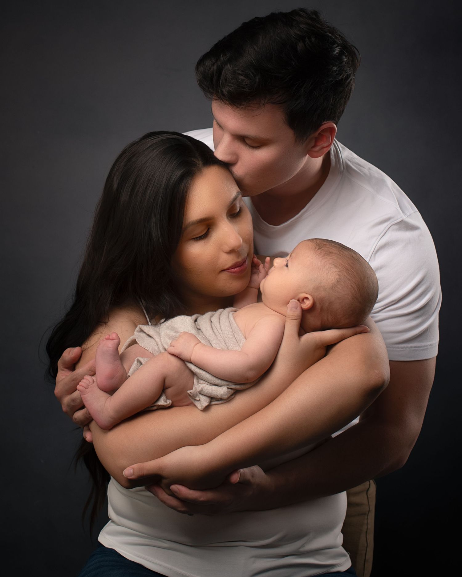 A couple tenderly holds their newborn baby, the man kissing the woman's head, in a professional studio portrait.