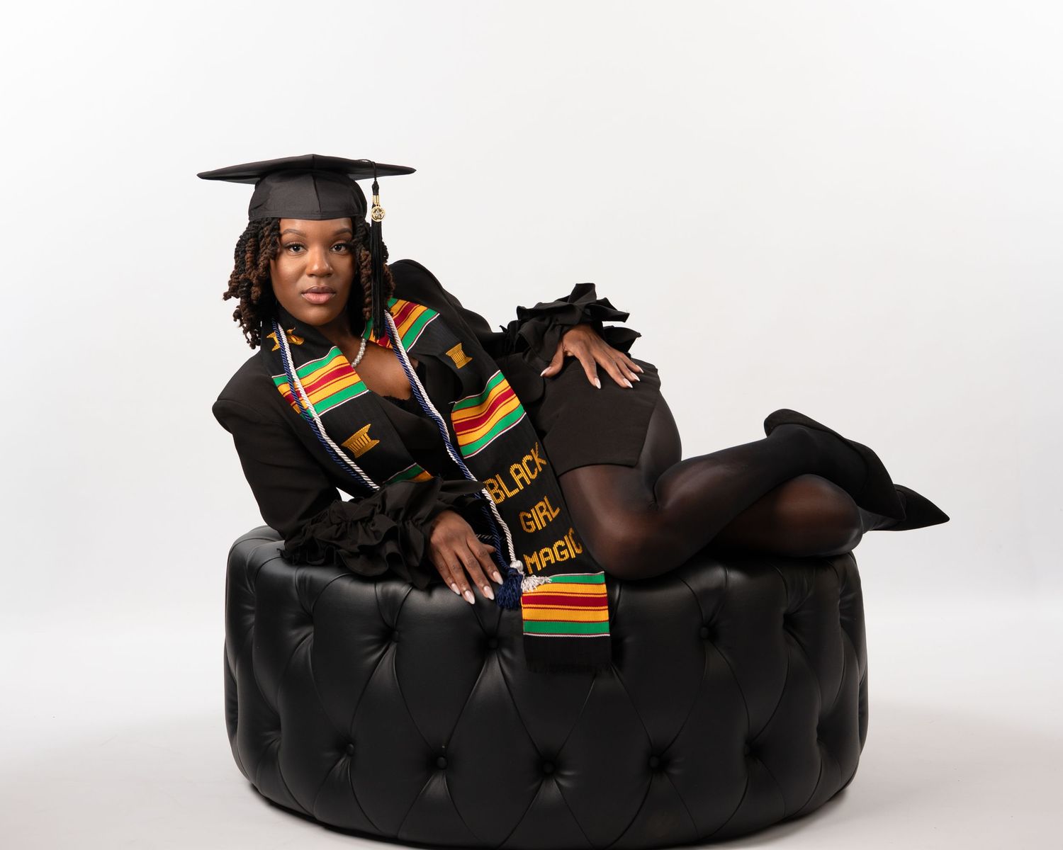 Graduate in cap and gown with Kente stole posing on tufted black ottoman.