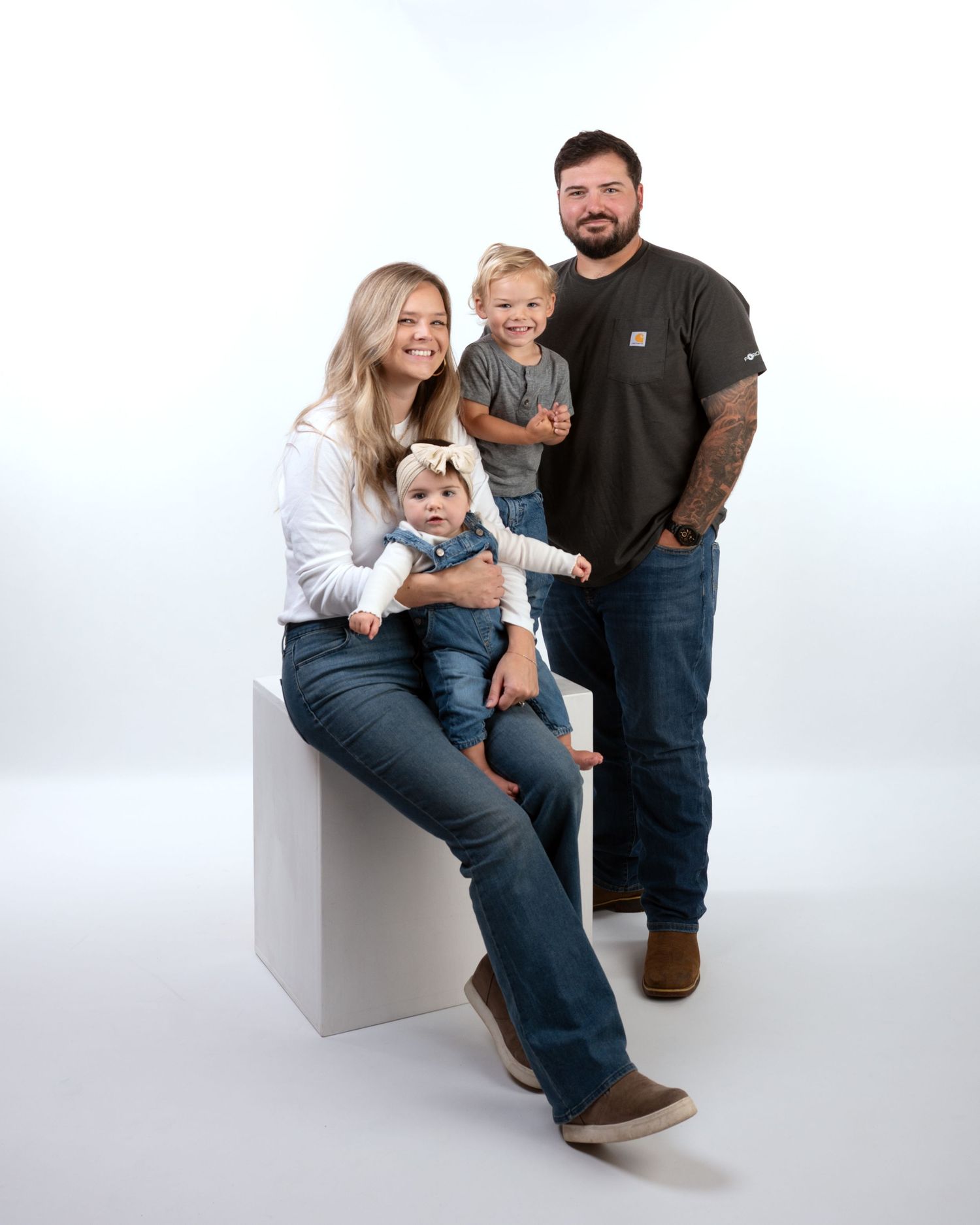 Young family of four with two small children posing on white cube in bright studio portrait.