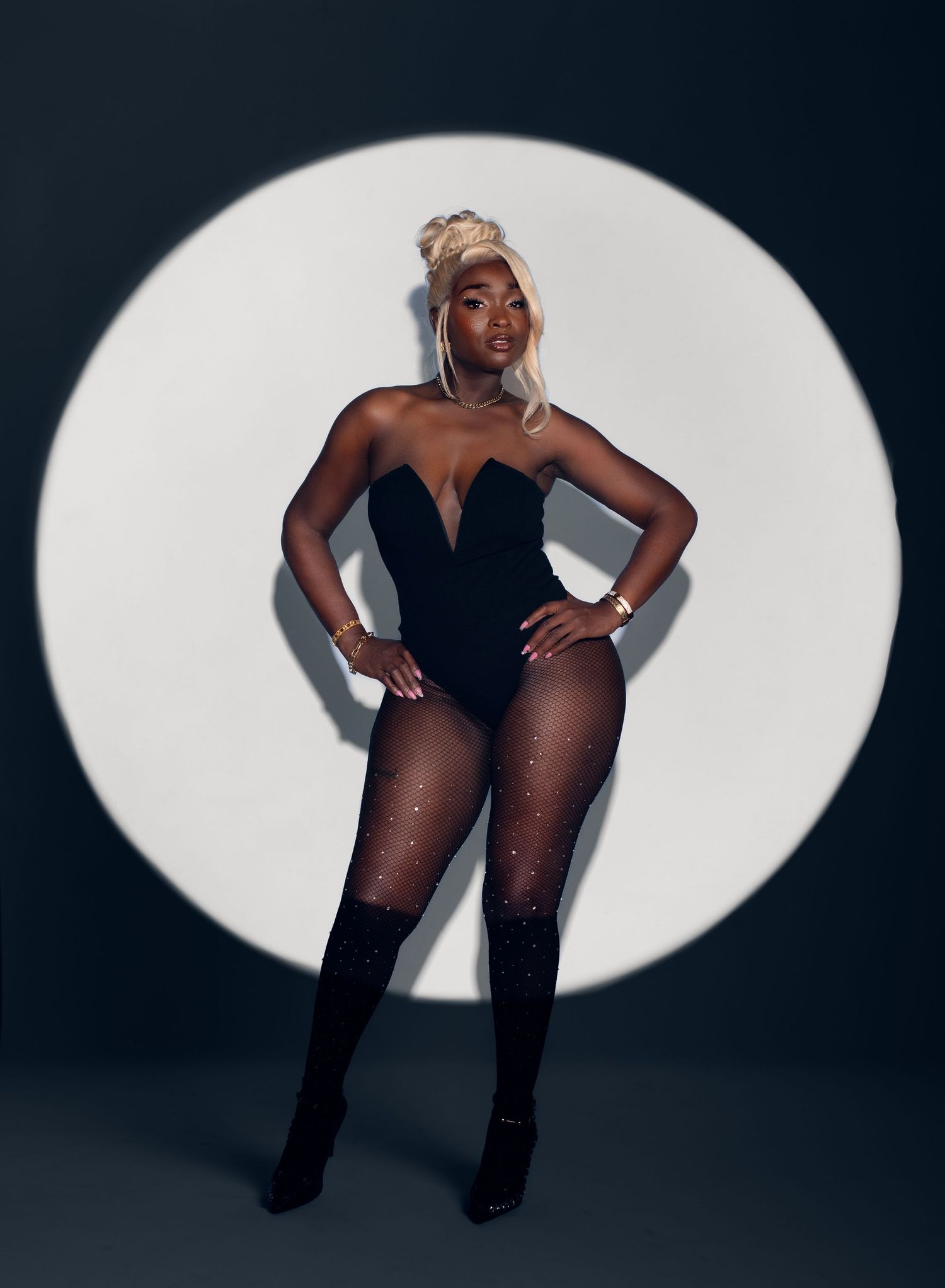Woman with blonde updo poses confidently in a black strapless bodysuit, sparkling tights and heeled boots against a bright spotlight circle.