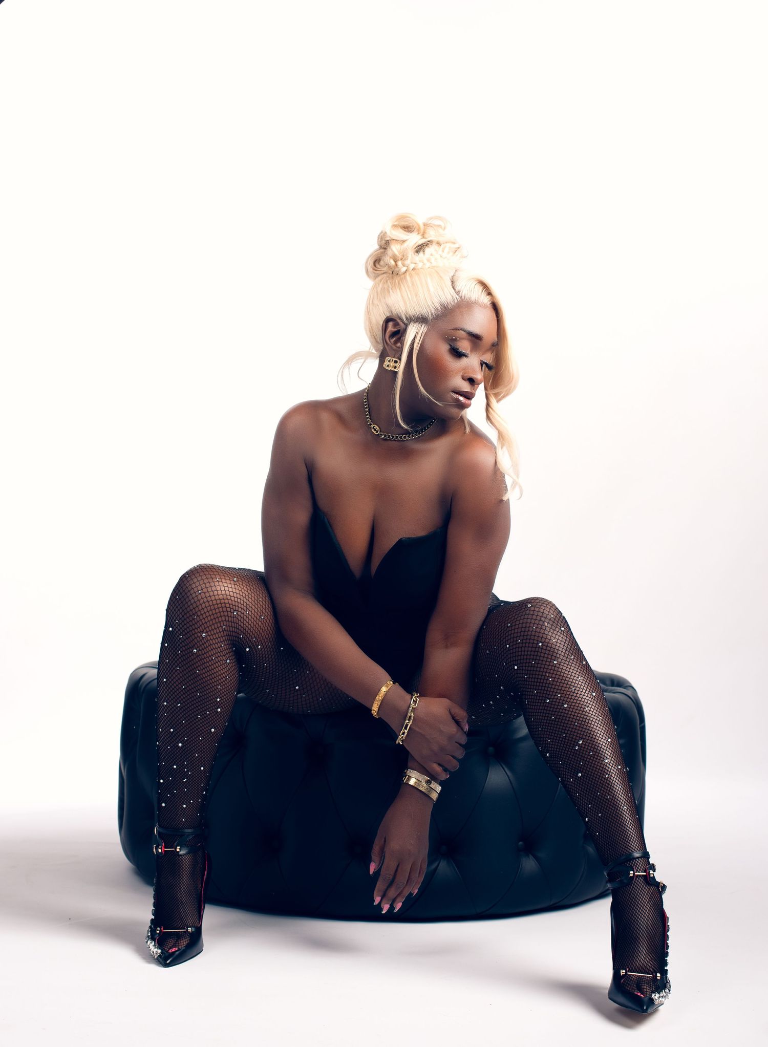 Woman with a blonde updo sits on a black tufted ottoman in a black bodysuit, sparkly tights and strappy heels against a white backdrop.