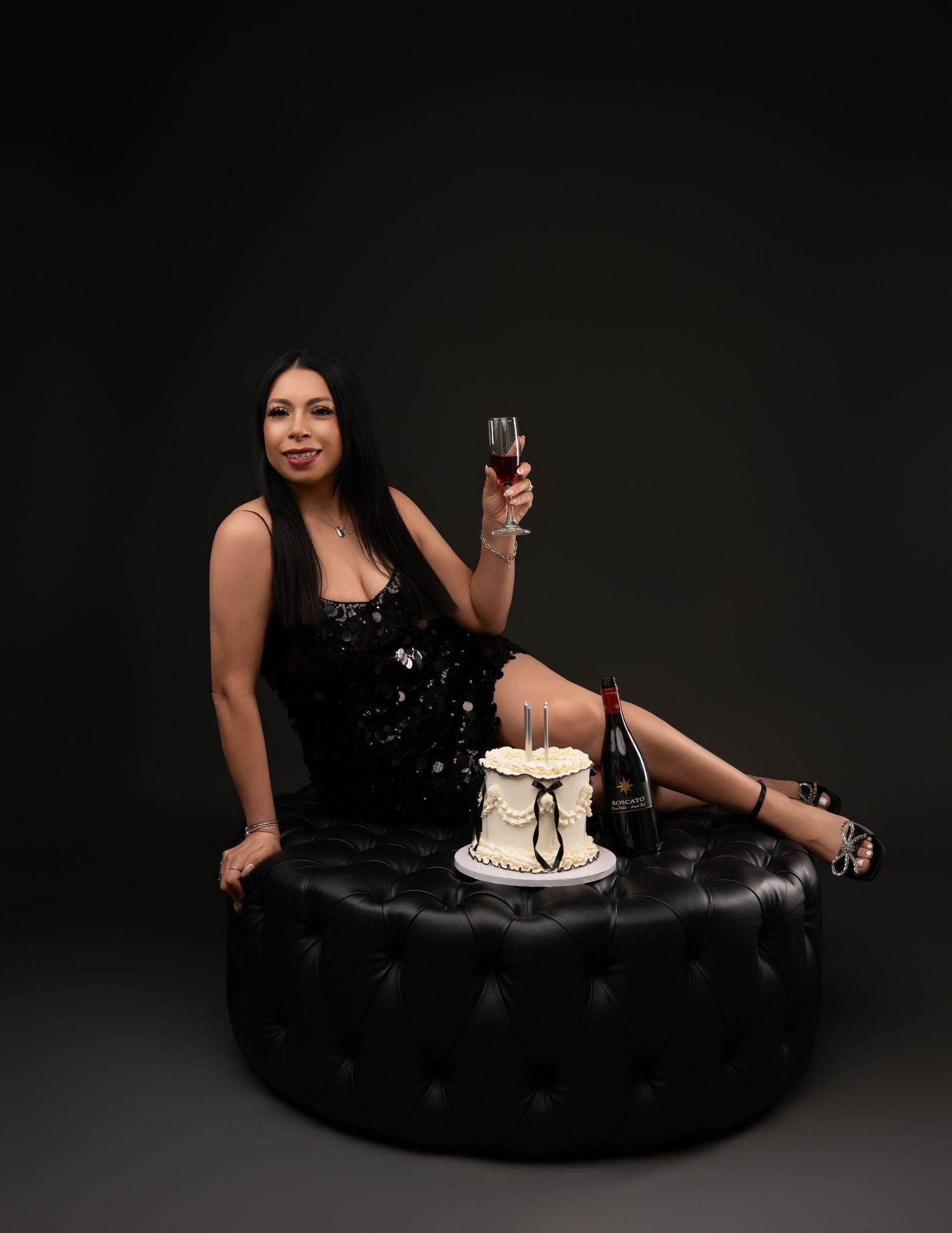 Woman in a black sequin dress reclines on a tufted black ottoman holding a wine glass beside a small white cake and a wine bottle.