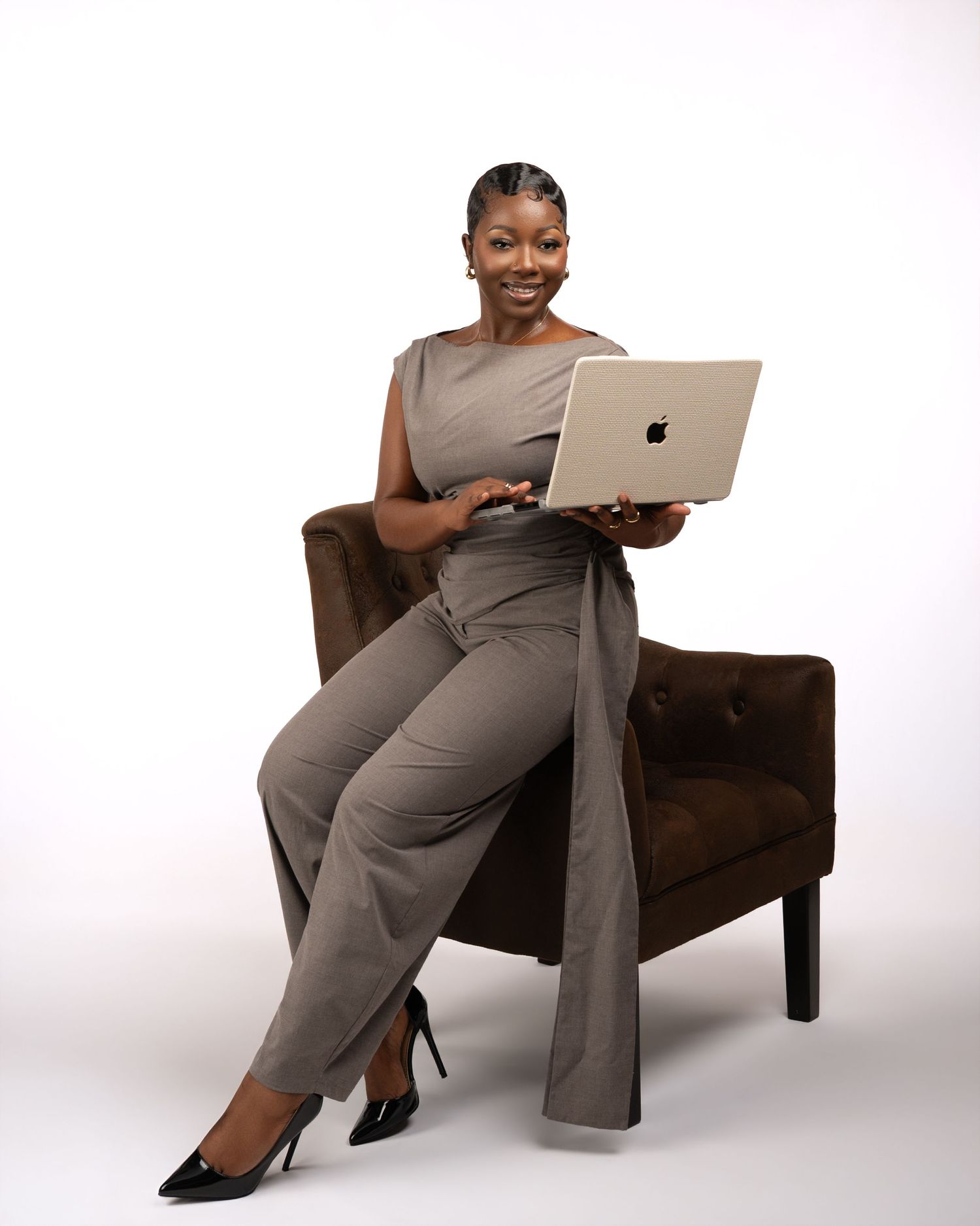 Professional woman in gray jumpsuit sitting in brown chair with MacBook laptop, smiling confidently.