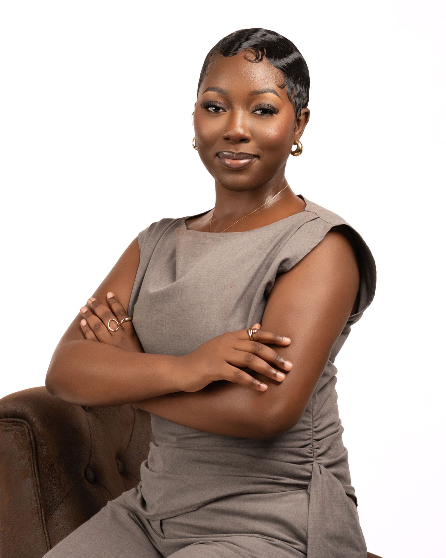 Professional woman in gray dress sitting confidently with arms crossed, wearing gold jewelry against white background.