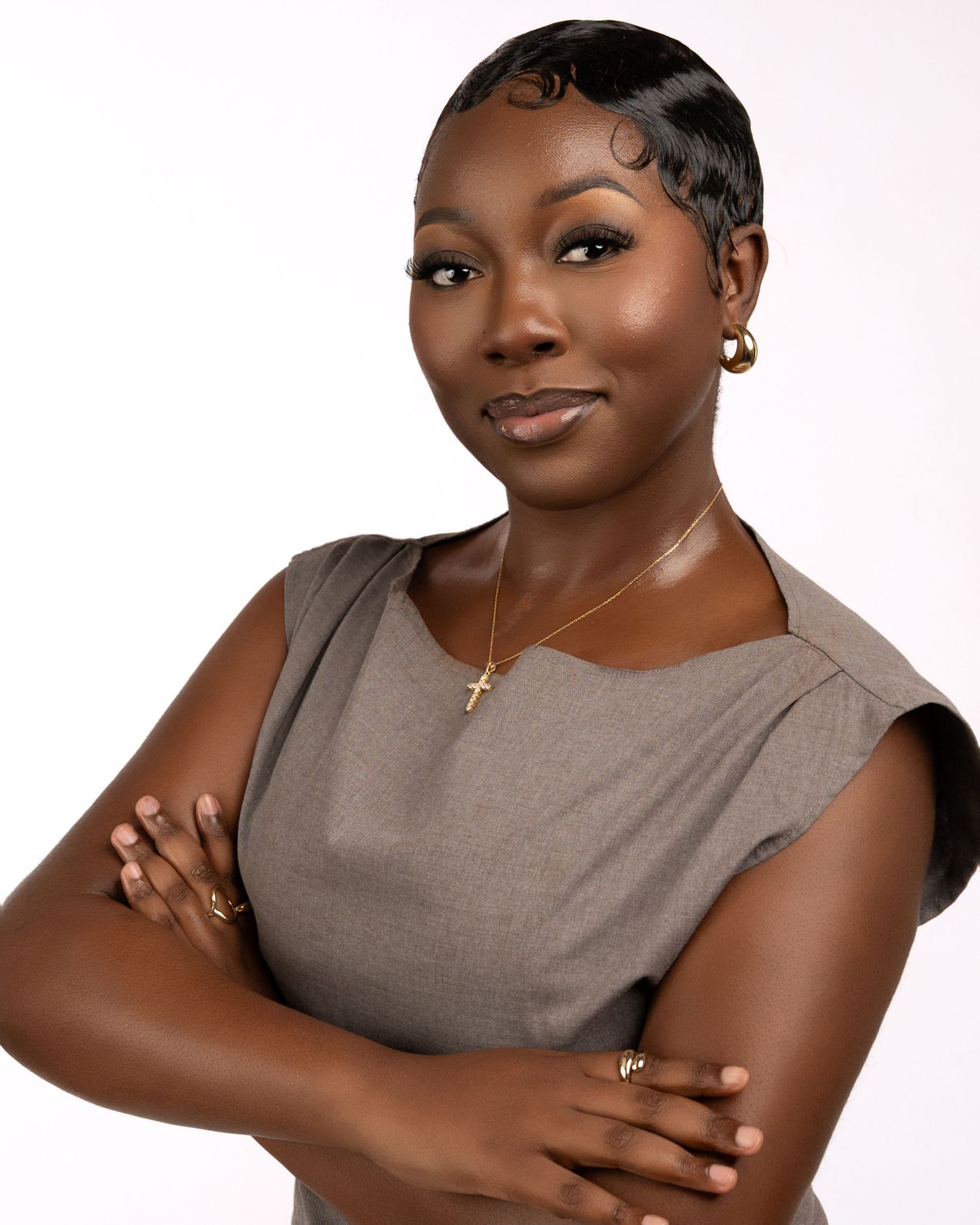 Professional woman in gray dress with cross necklace, arms crossed, posing confidently against white background.
