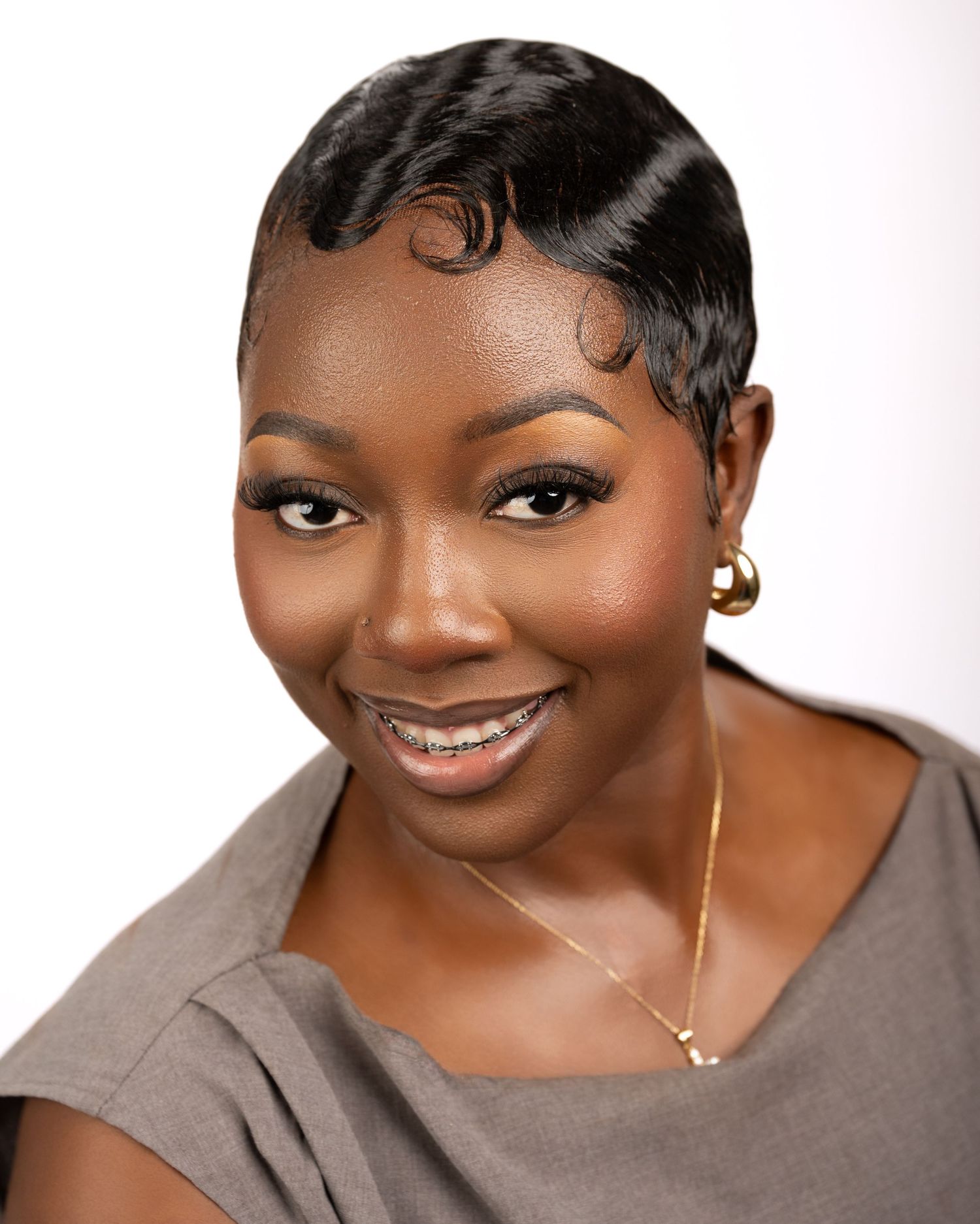 Professional headshot of a smiling woman wearing gold earrings, necklace, and a gray top against a white background.