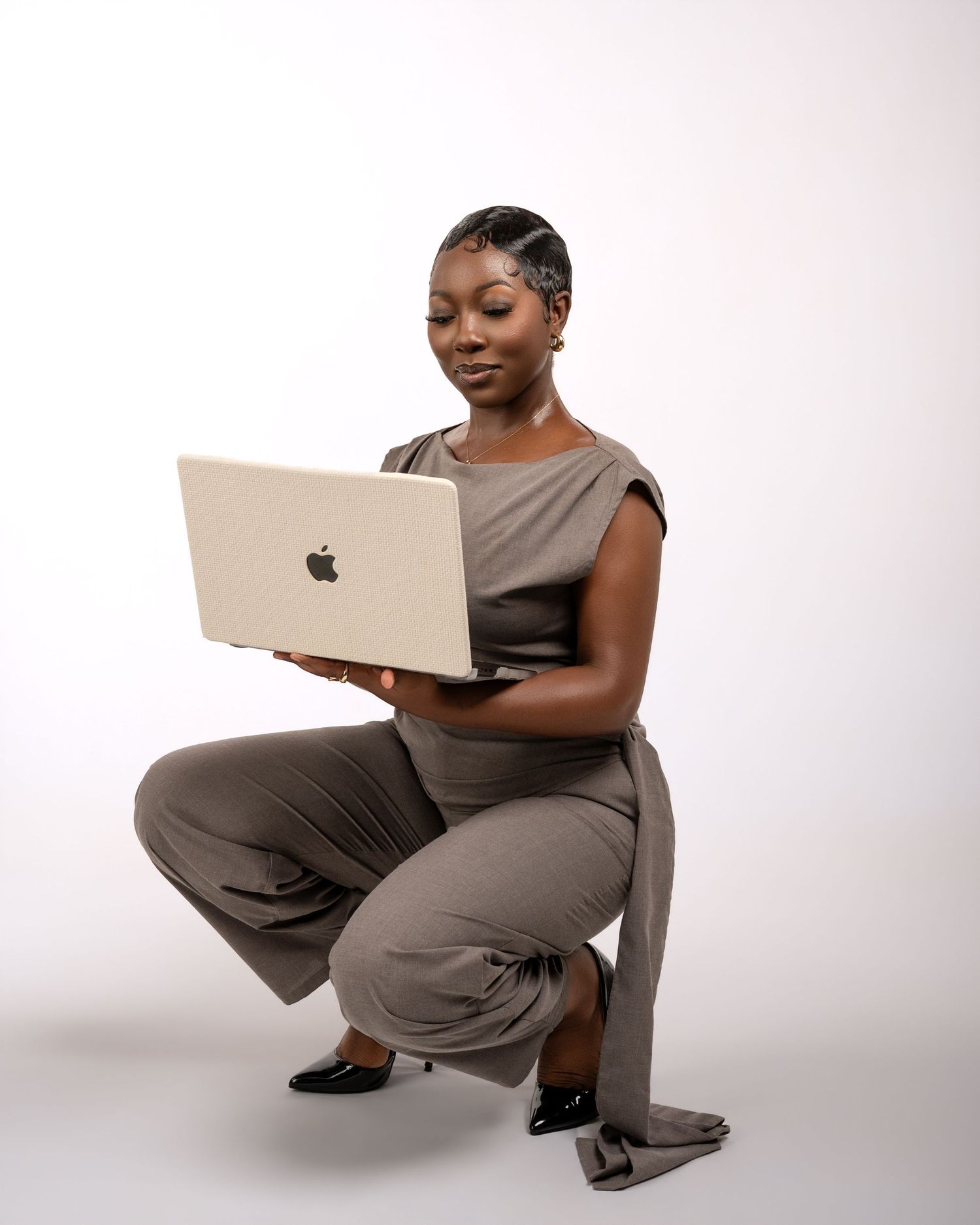 A professional Black woman in a gray jumpsuit crouches while holding an Apple MacBook laptop against a white background.