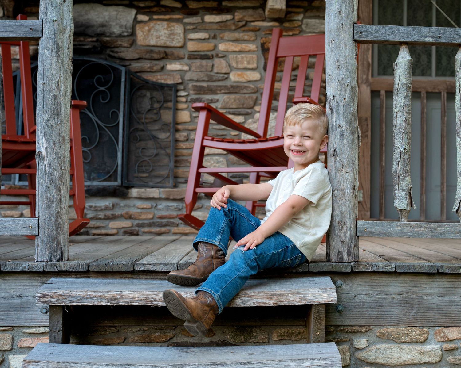 A cheerful young boy sits smiling on wooden porch steps of a rustic stone cabin.