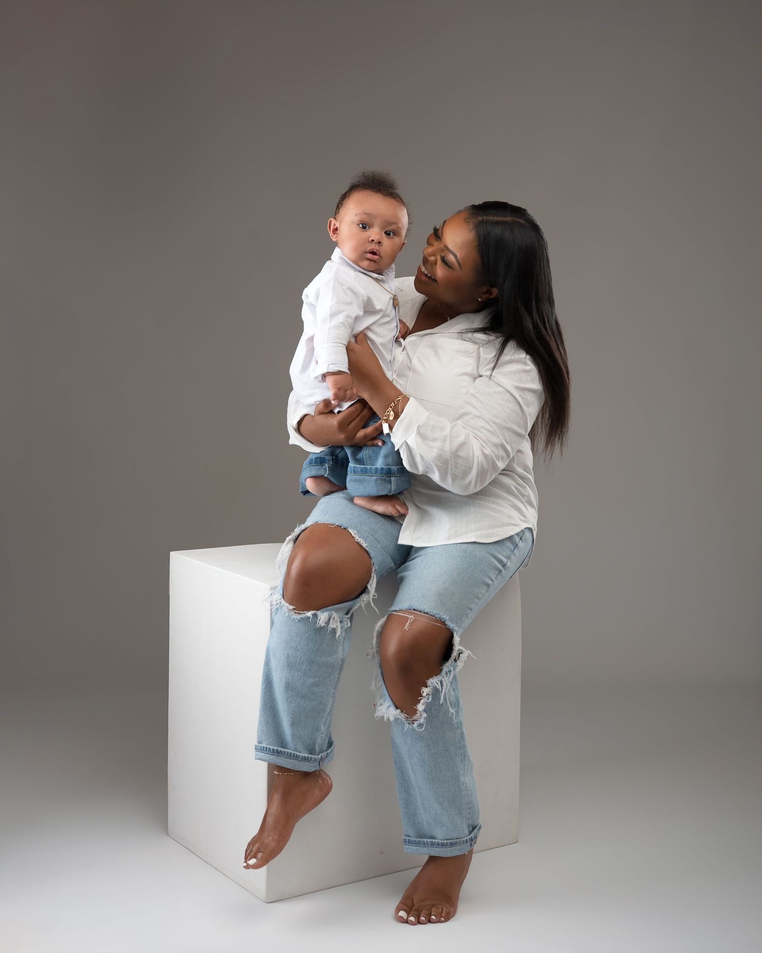 Mother in white shirt and ripped jeans holds smiling baby boy during studio portrait session.