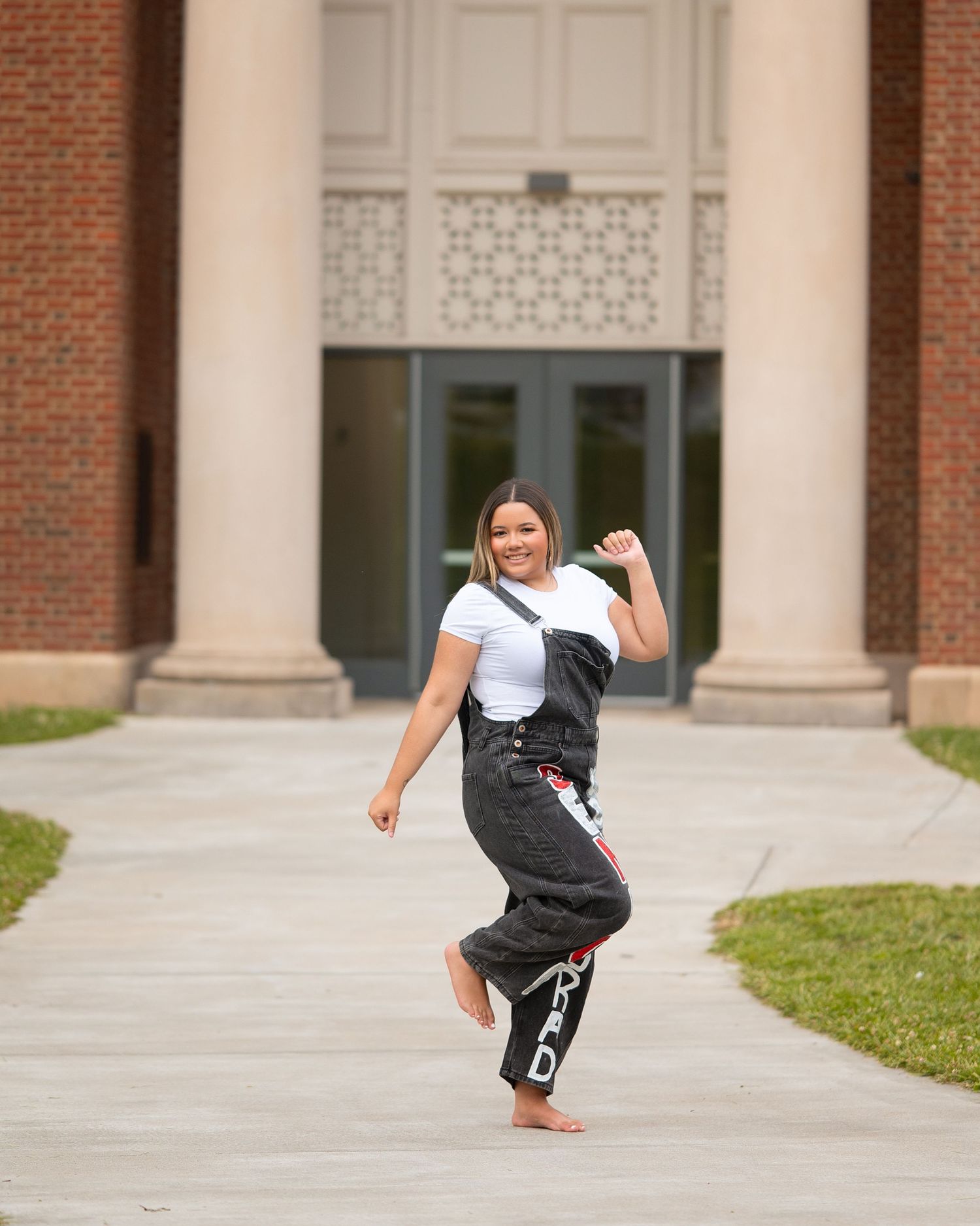 Happy young woman dances barefoot on campus walkway wearing gray sweatpants and white top with crossbody bag.