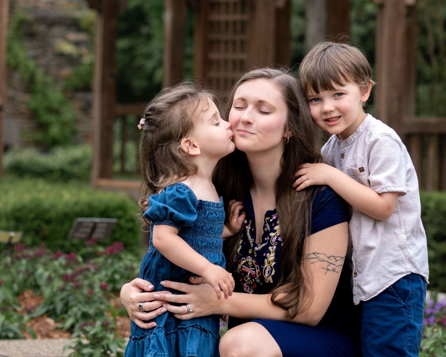Mother sitting outdoors with two young children hugging and kissing her cheeks affectionately.