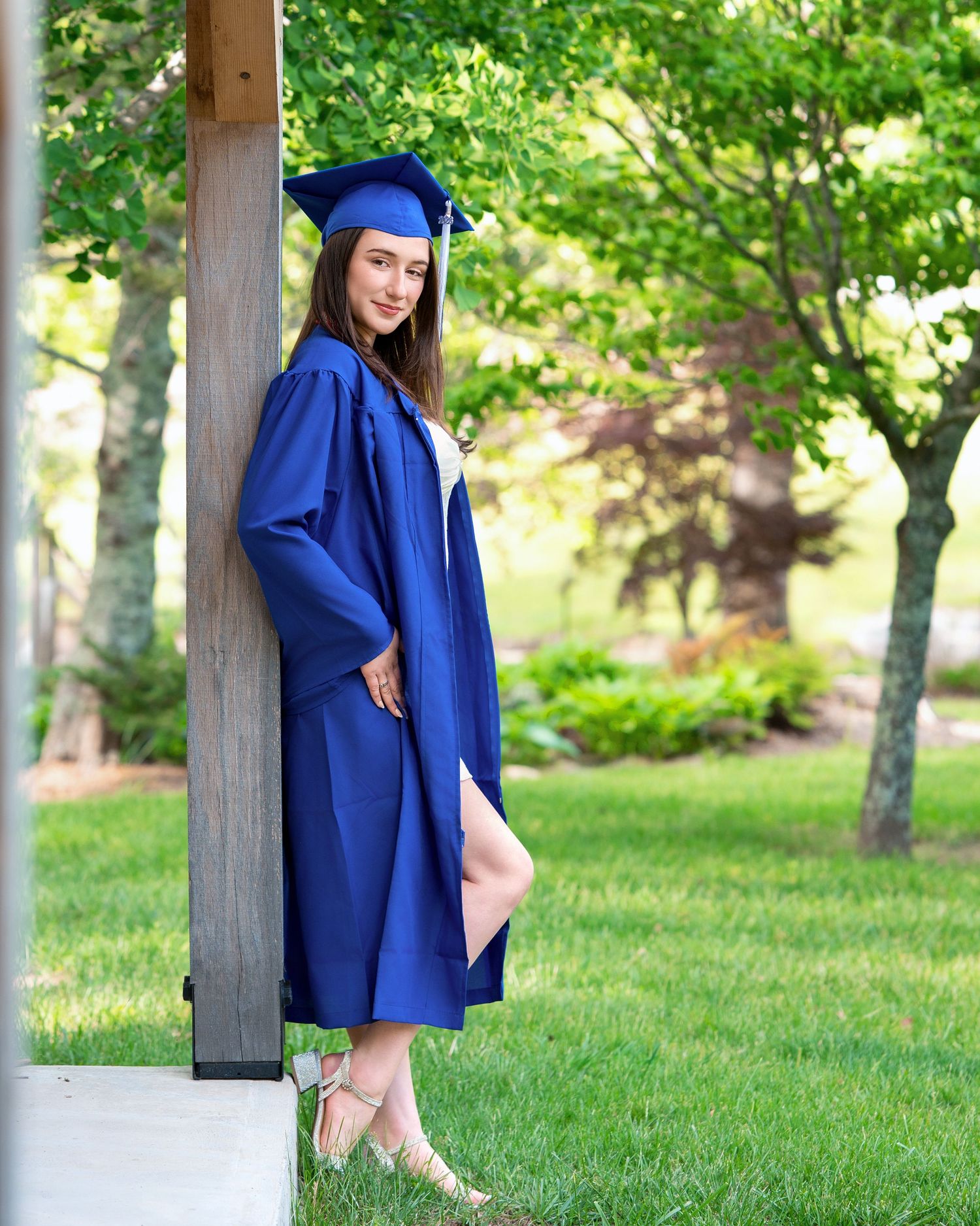 Young woman in blue graduation cap and gown leans against wooden post in green garden.