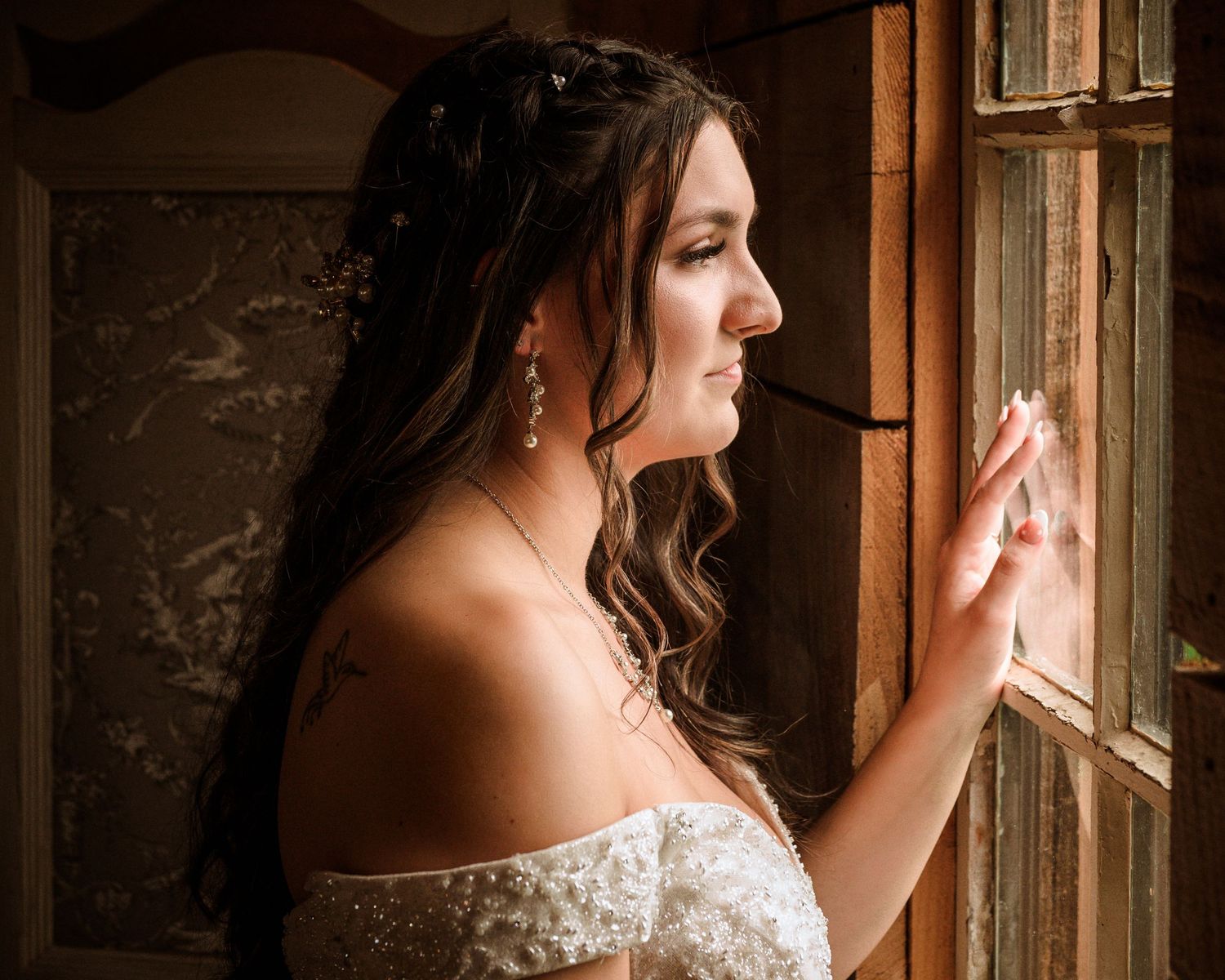 Beautiful bride in a lace off-shoulder gown gazing out a window in soft dramatic lighting.