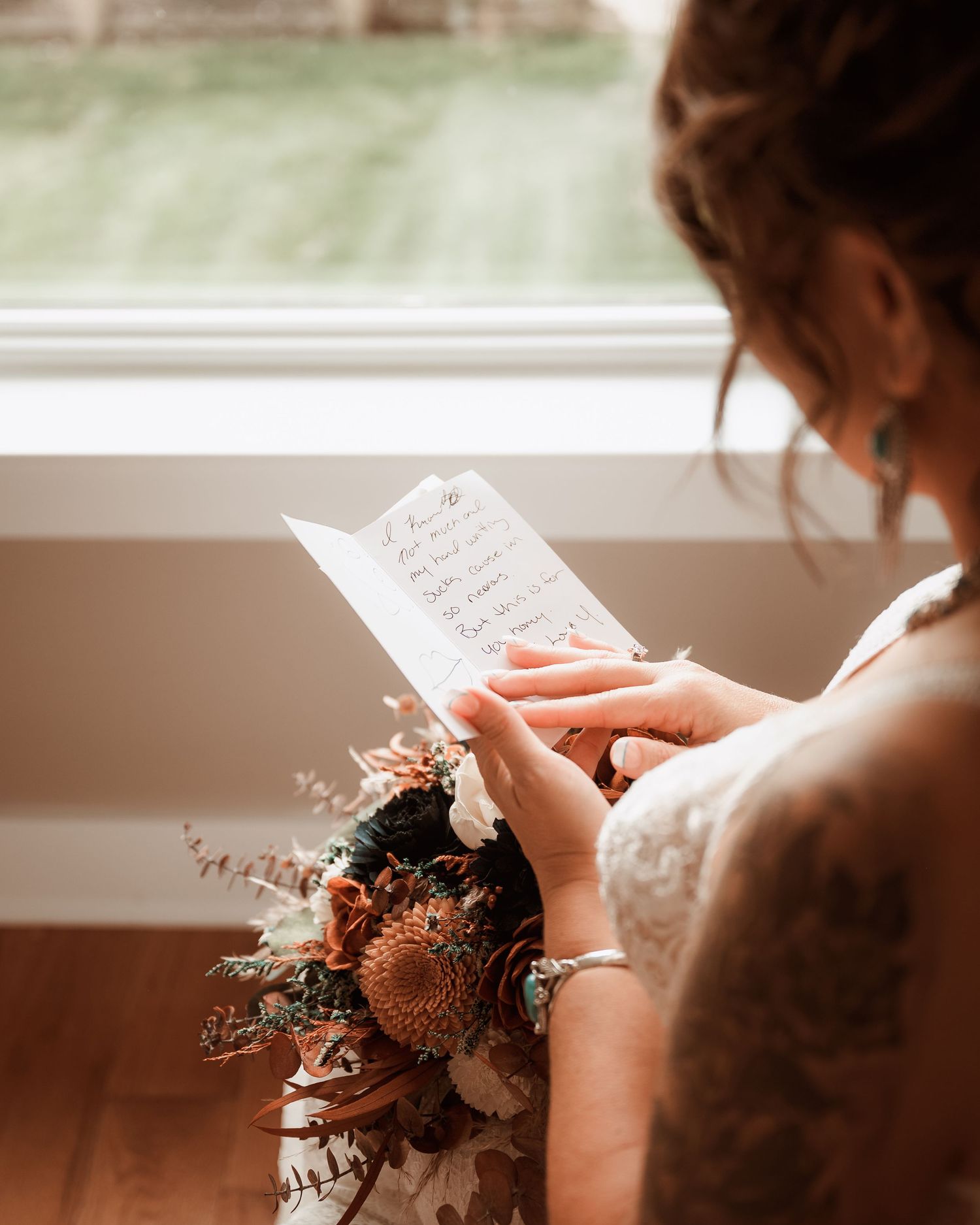 Bride in lace gown reads a heartfelt letter while holding a rustic bouquet near a sunlit window.
