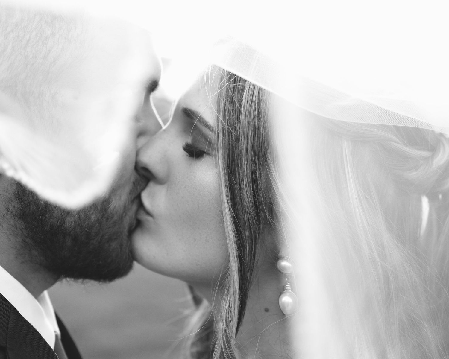 Black and white intimate close-up of bride and groom kissing beneath flowing wedding veil.
