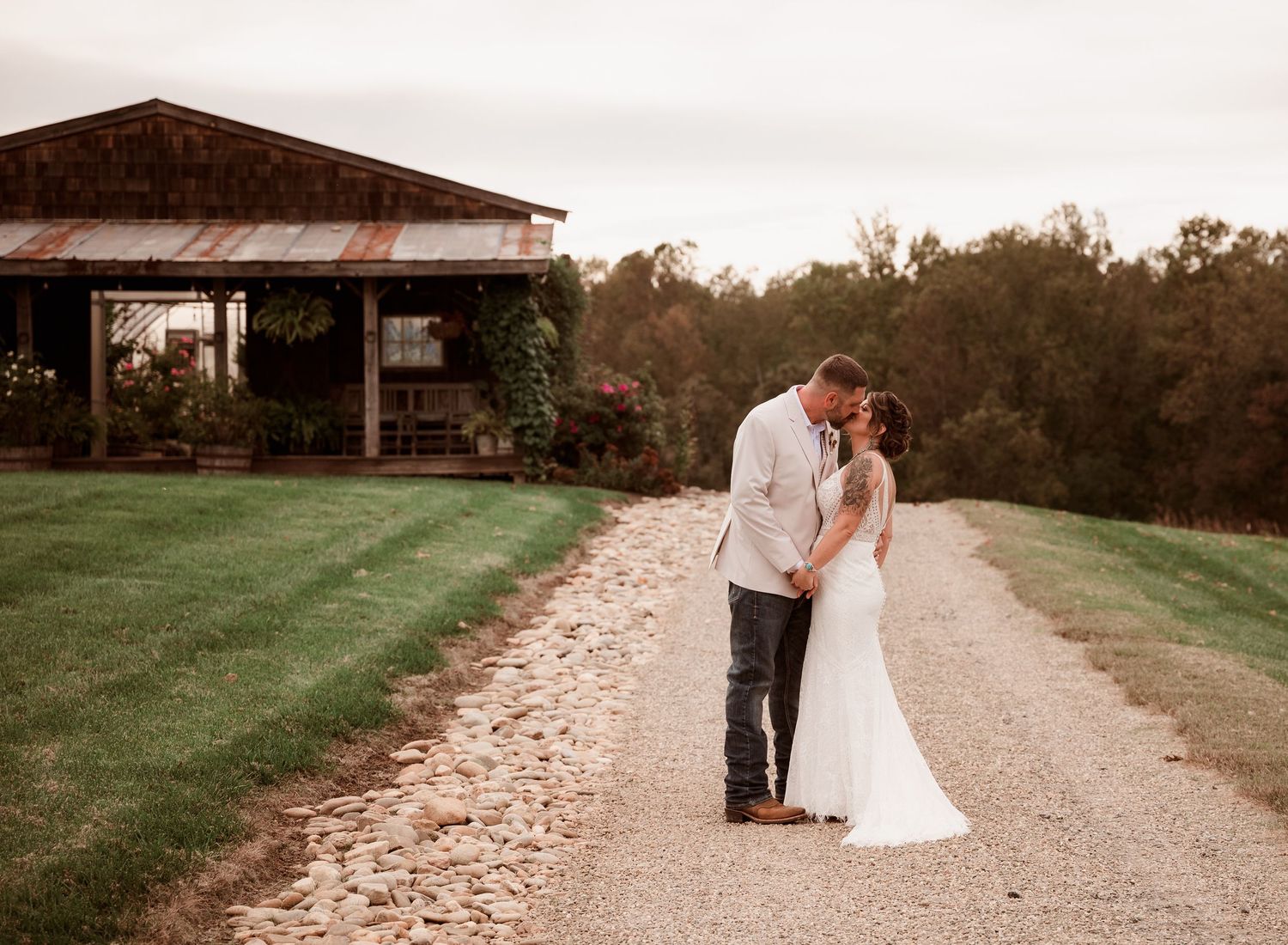Bride and groom share a kiss on a gravel path near a rustic barn at dusk.