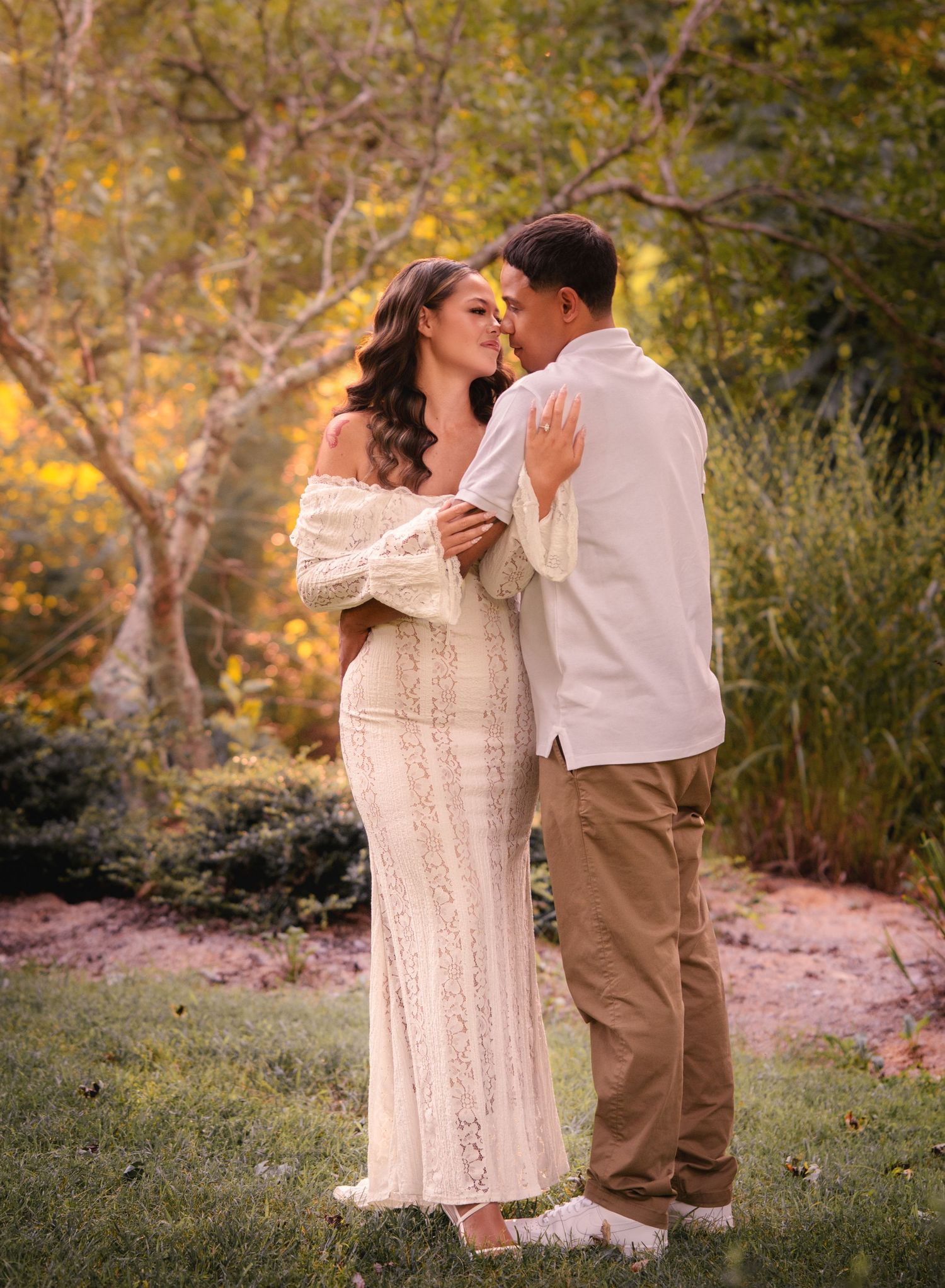 Couple nearly kissing in lush garden setting, woman in white lace gown and man in white shirt and khakis.