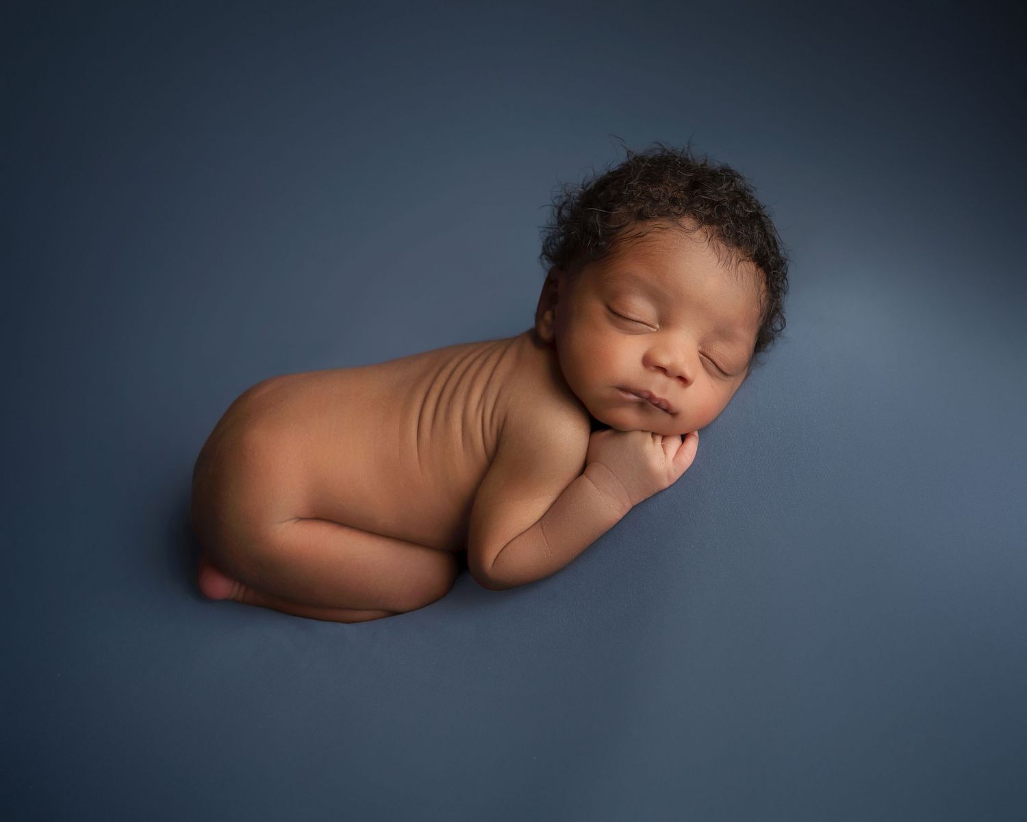 Peaceful newborn baby sleeping curled up on a blue backdrop in a professional studio portrait.