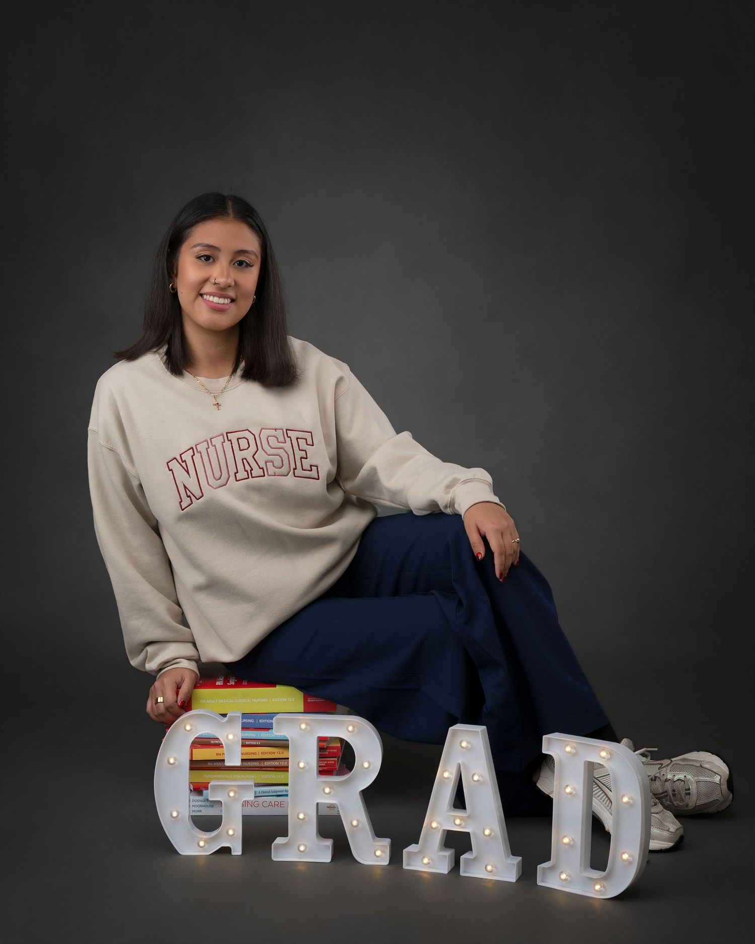 Nursing graduate smiles wearing a beige NURSE sweatshirt beside lit GRAD letters and textbooks.