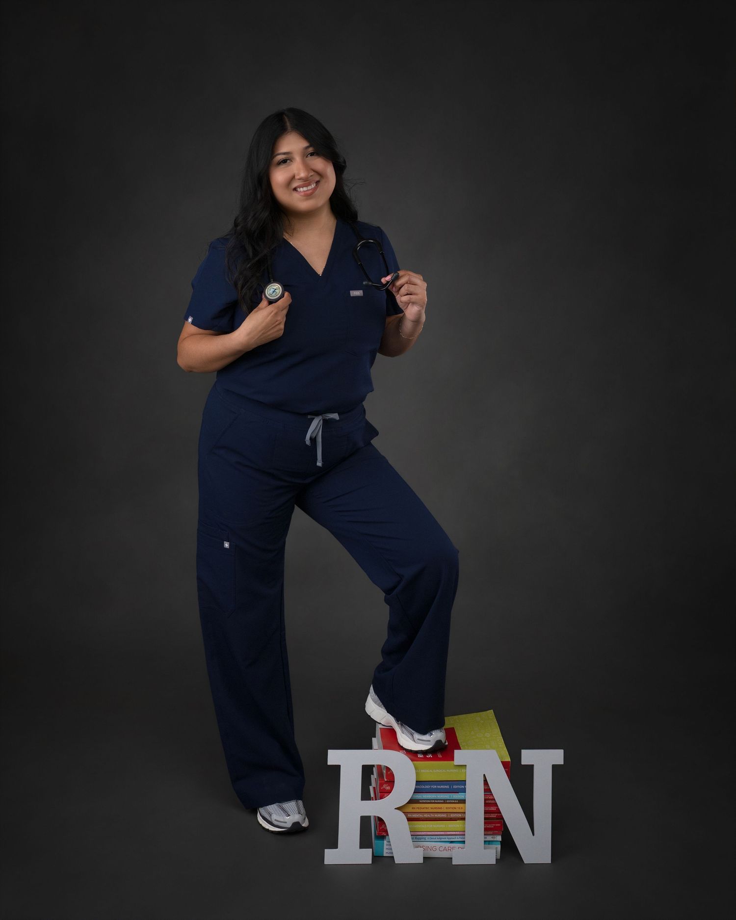 Proud new RN poses in navy scrubs with stethoscope standing on letter blocks in studio.