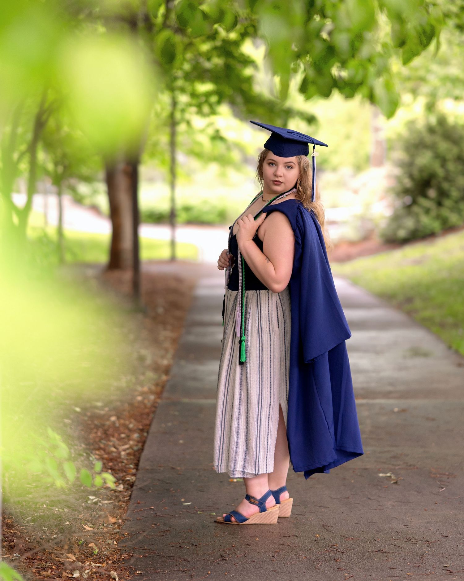 Female graduate in blue cap and gown holding regalia on a tree-lined campus walkway, looking over shoulder.