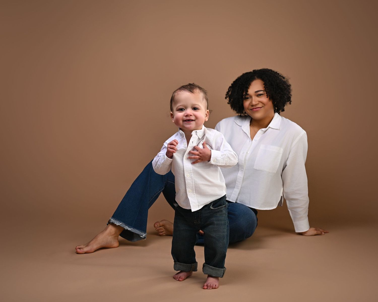 A smiling mother in white shirt and jeans poses with her standing toddler on a tan background.