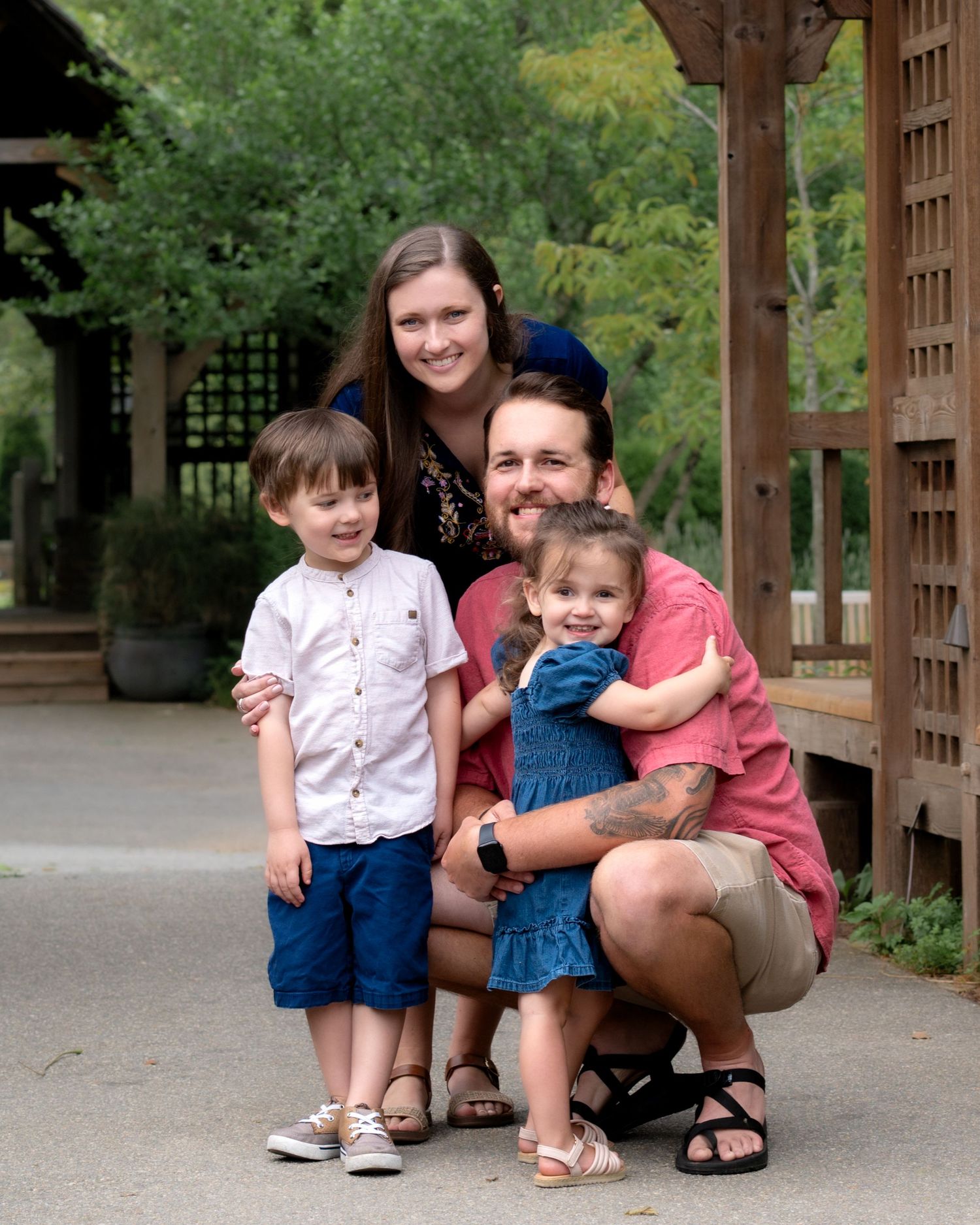 Happy family of four posing together under a wooden pergola surrounded by green trees.