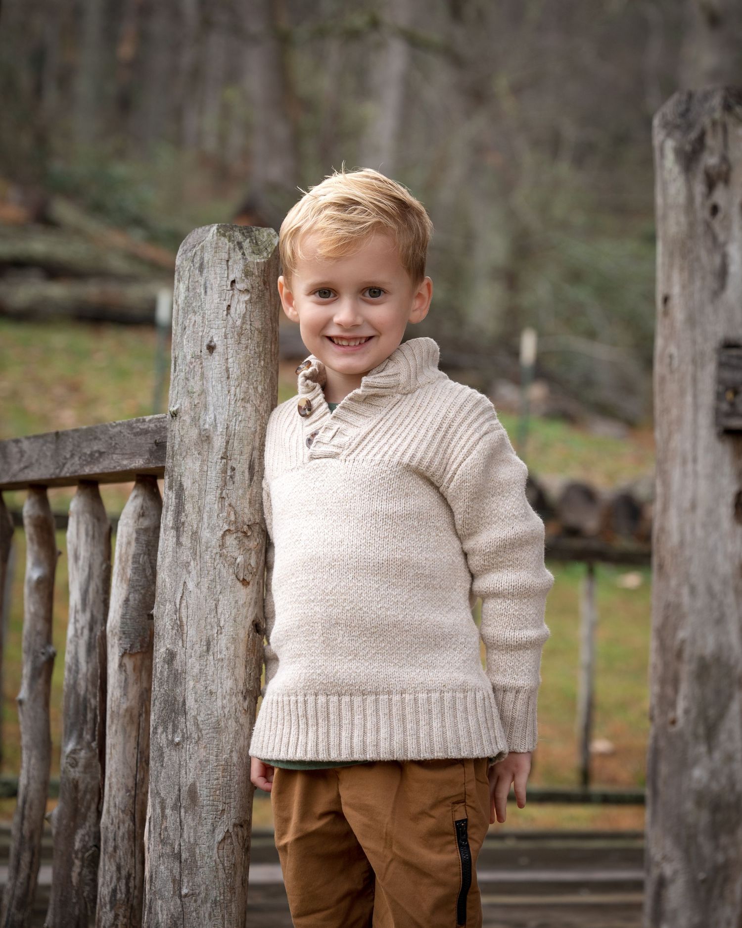 Smiling young boy in cream knit sweater leaning against rustic wooden fence in outdoor nature setting.