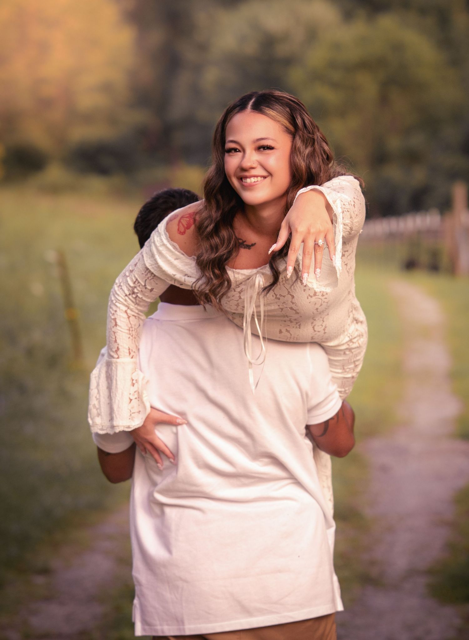 Smiling woman in white lace dress showing engagement ring while getting a piggyback ride outdoors.