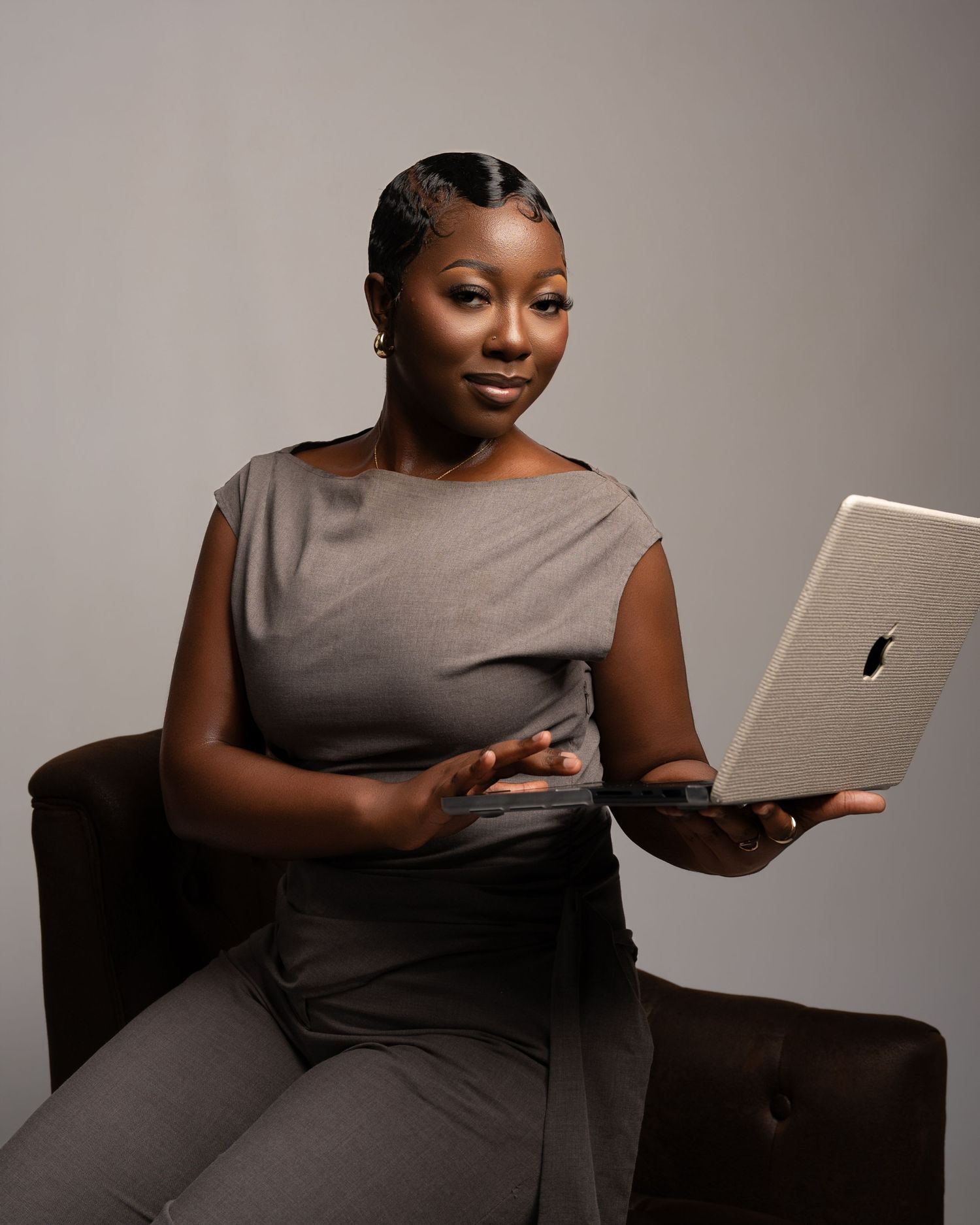 Professional woman in gray outfit holding Apple MacBook laptop while seated in a dark chair against neutral background.