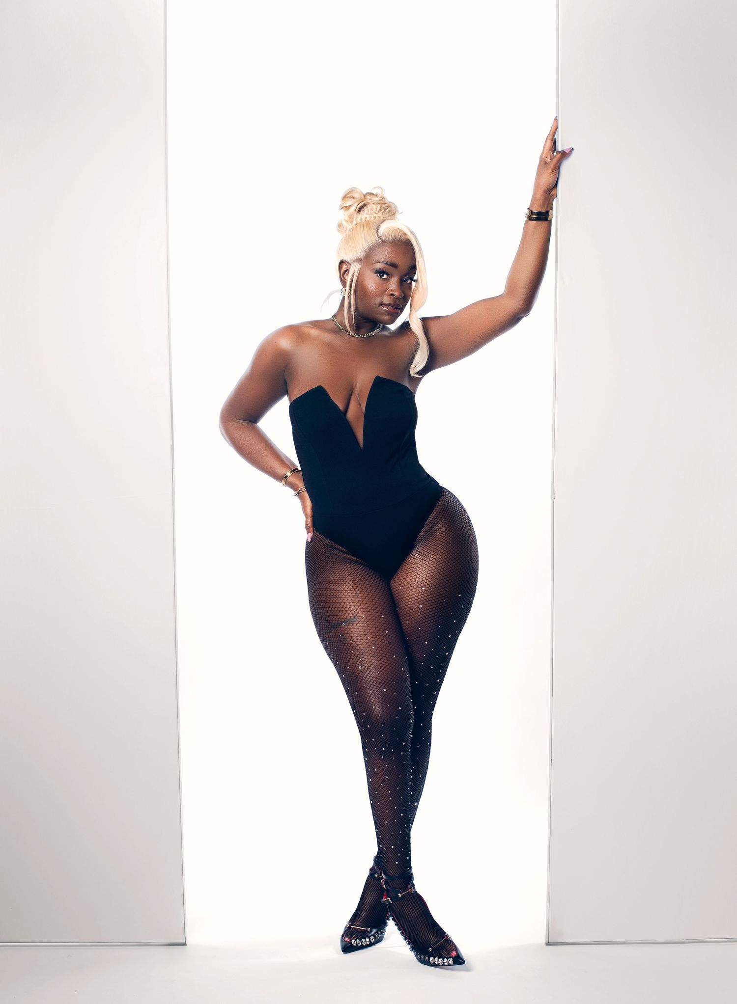 Woman with a blonde updo poses against a white doorway in a black plunging bodysuit, sparkly tights and embellished heels.