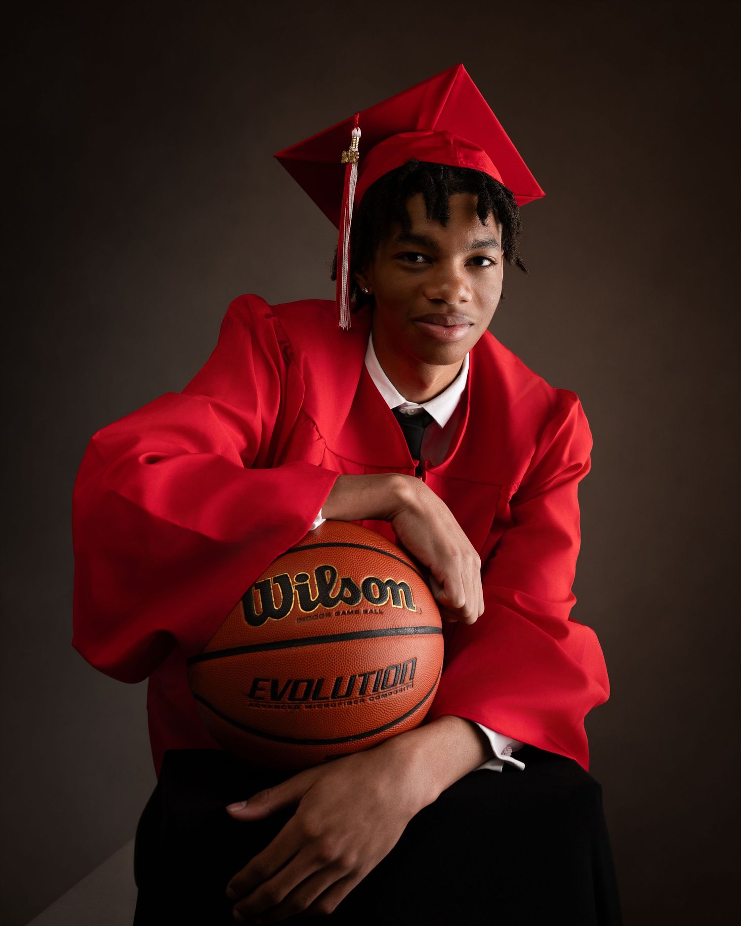 Young male graduate in red cap and gown poses with a Wilson basketball in a studio portrait.