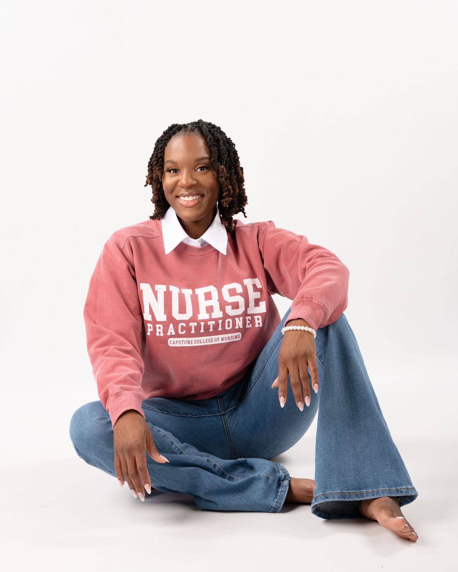 Smiling woman in pink Nurse Practitioner sweatshirt sitting cross-legged on white floor.