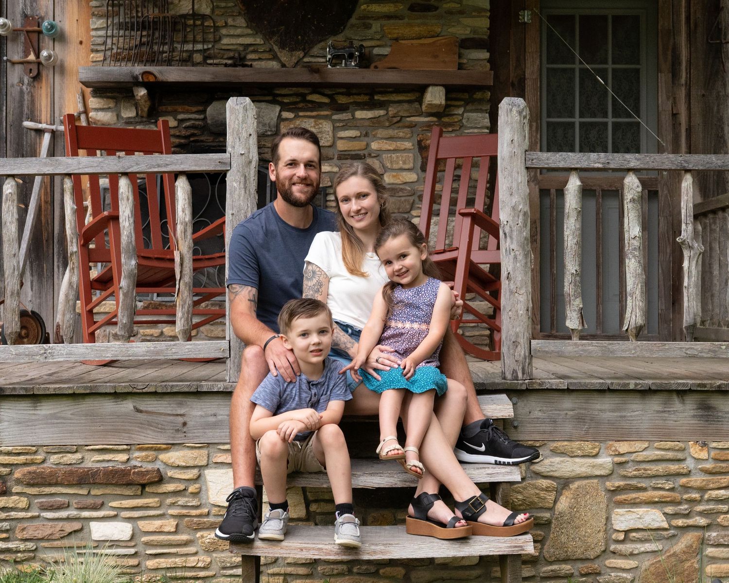 A smiling family of four sits together on stone steps of a charming rustic log cabin porch.