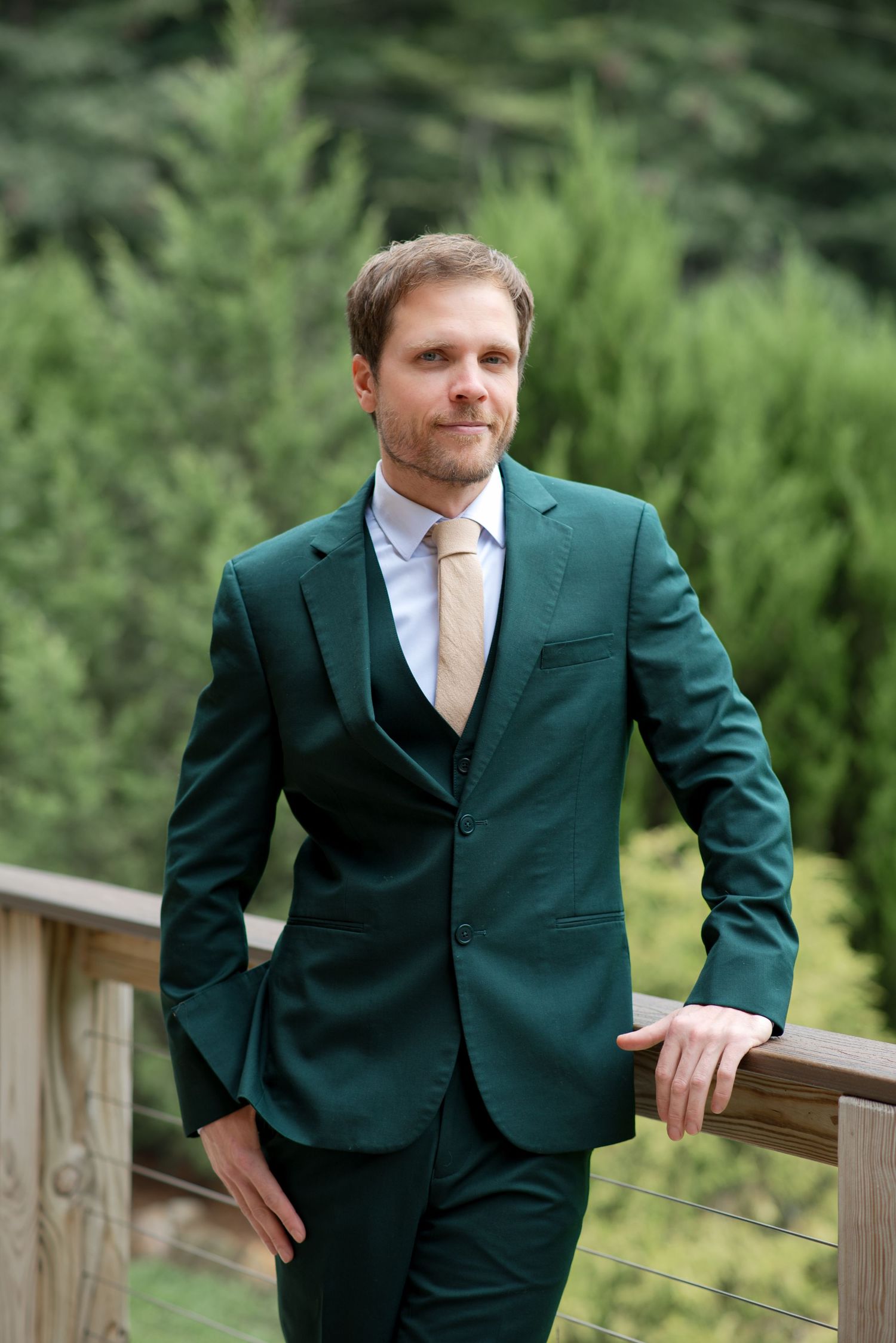 Stylish groom in emerald green suit leaning on wooden railing surrounded by pine trees.