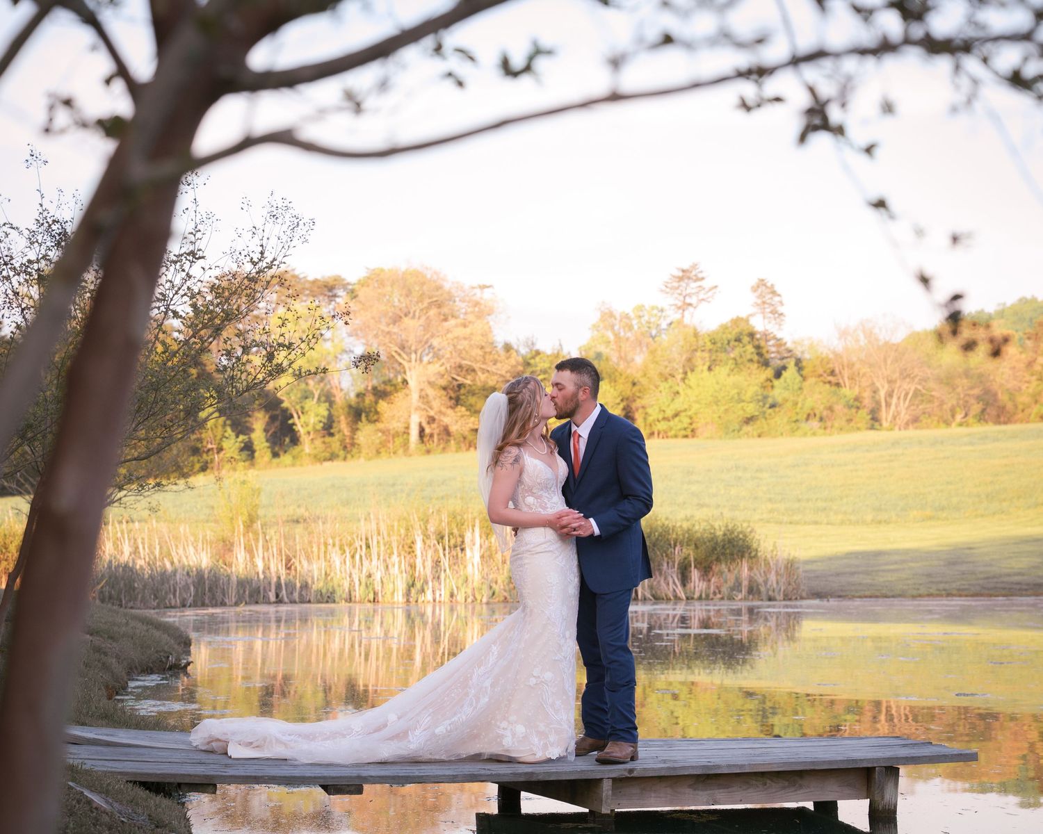 Bride and groom sharing a romantic kiss on a wooden dock by a pond at golden hour sunset.