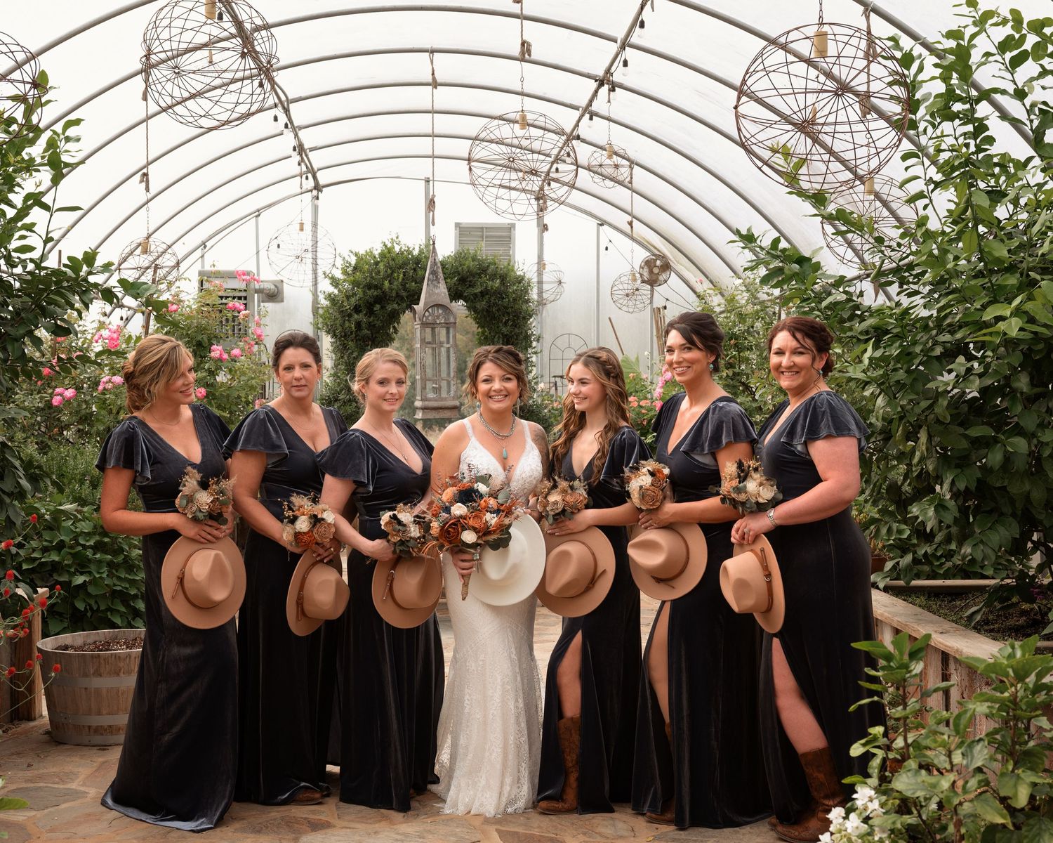 Wedding party in navy gowns with straw hats posing with bride in white lace dress in greenhouse.