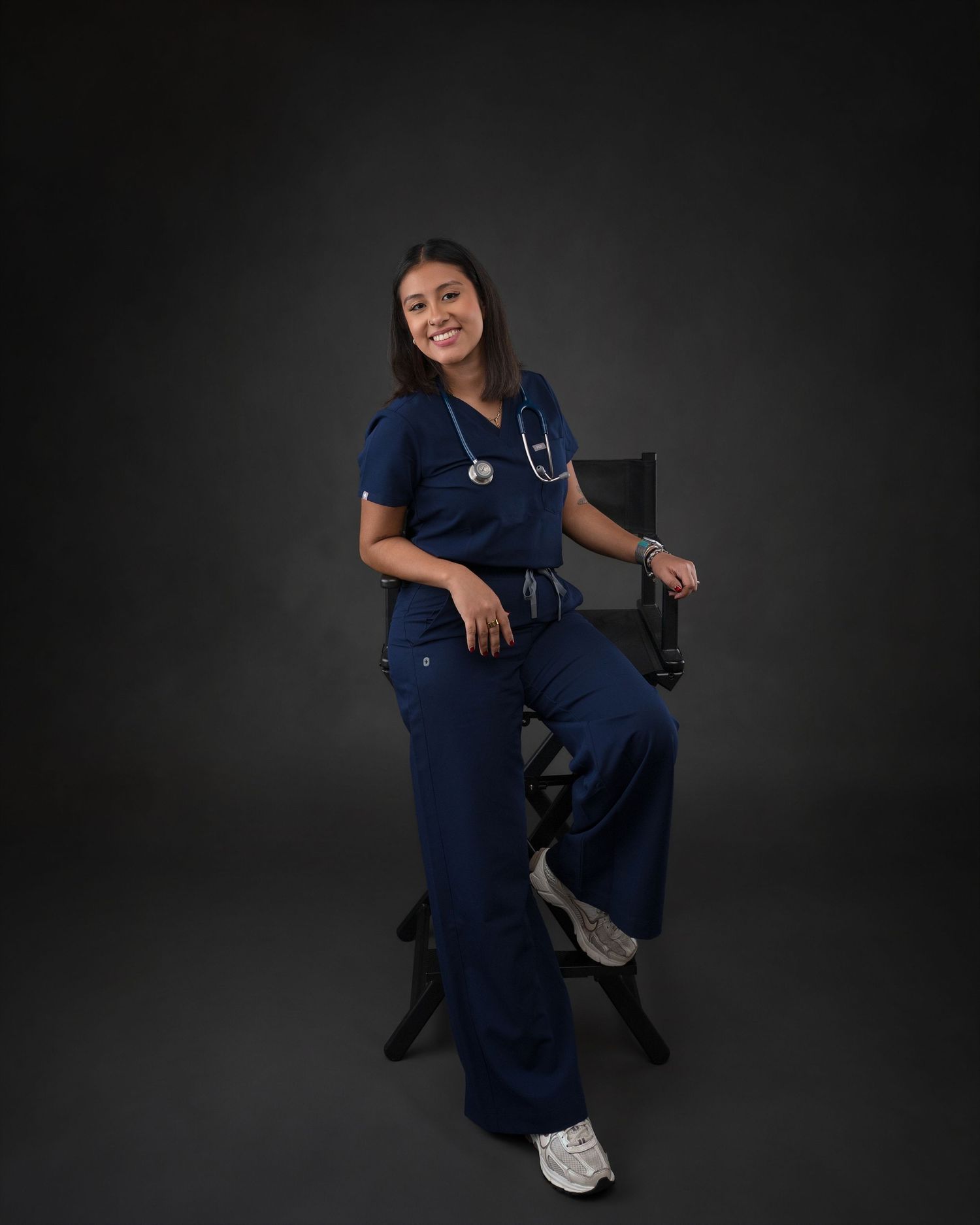 Smiling woman in navy blue scrubs with stethoscope seated on chair for nursing graduation portrait.