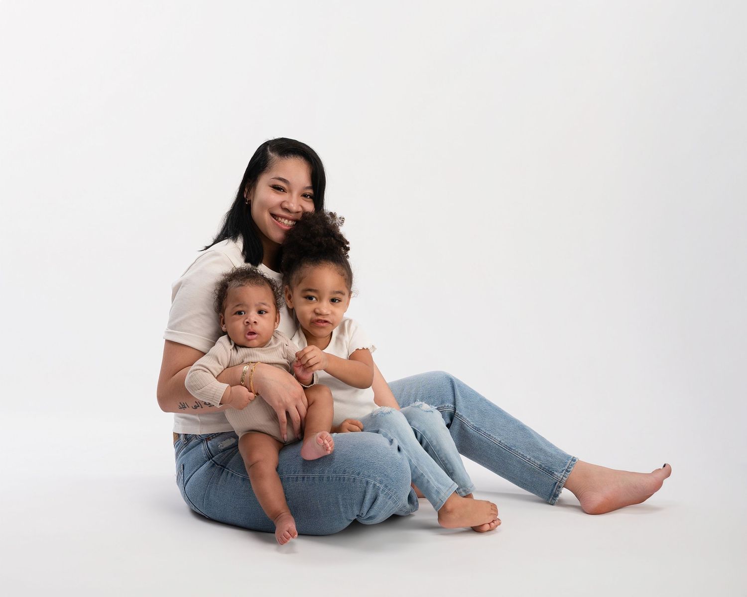 Smiling mother sits on floor holding two young children during bright white studio family portrait.