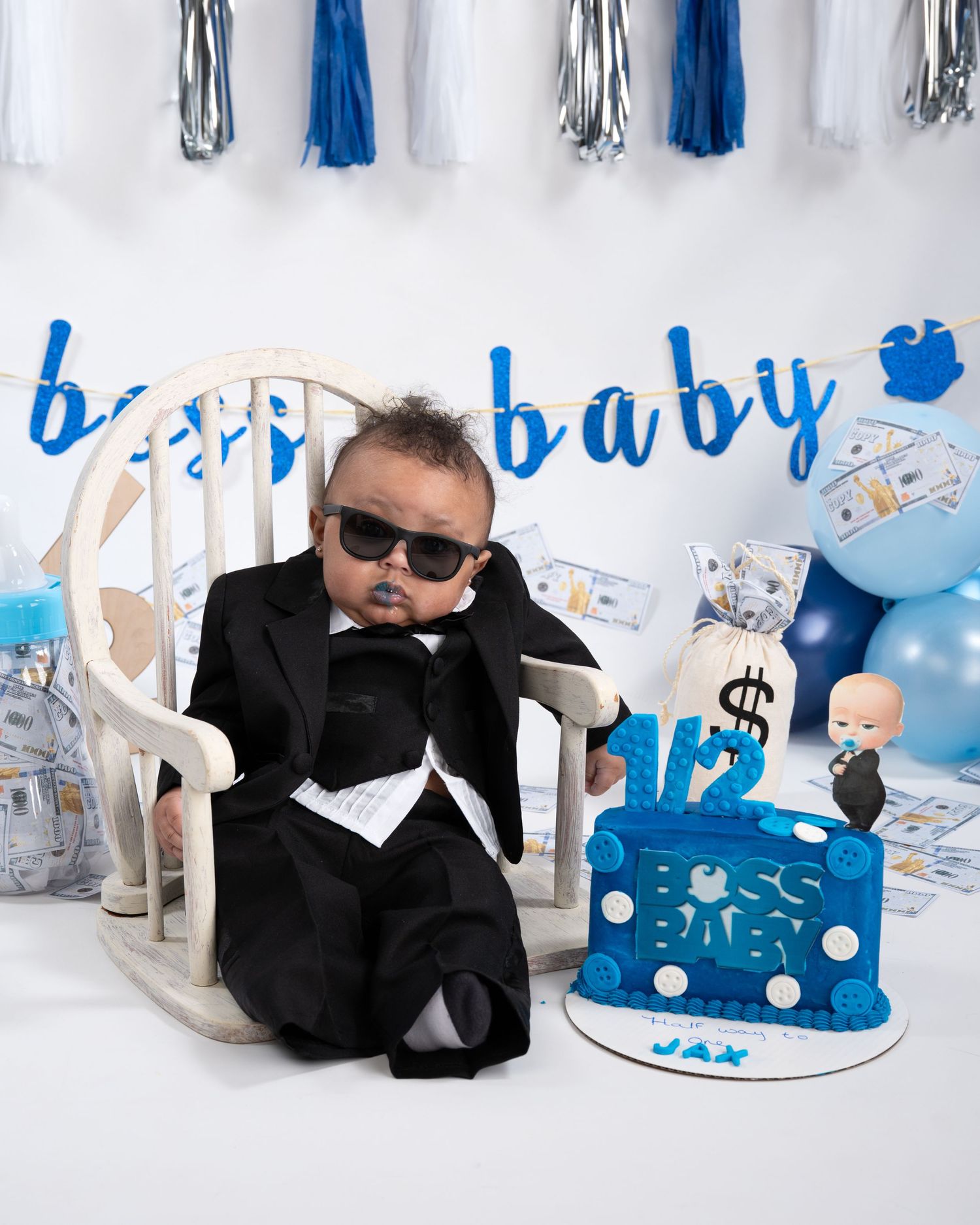 Baby boy in black suit and sunglasses poses beside boss baby themed half birthday cake.