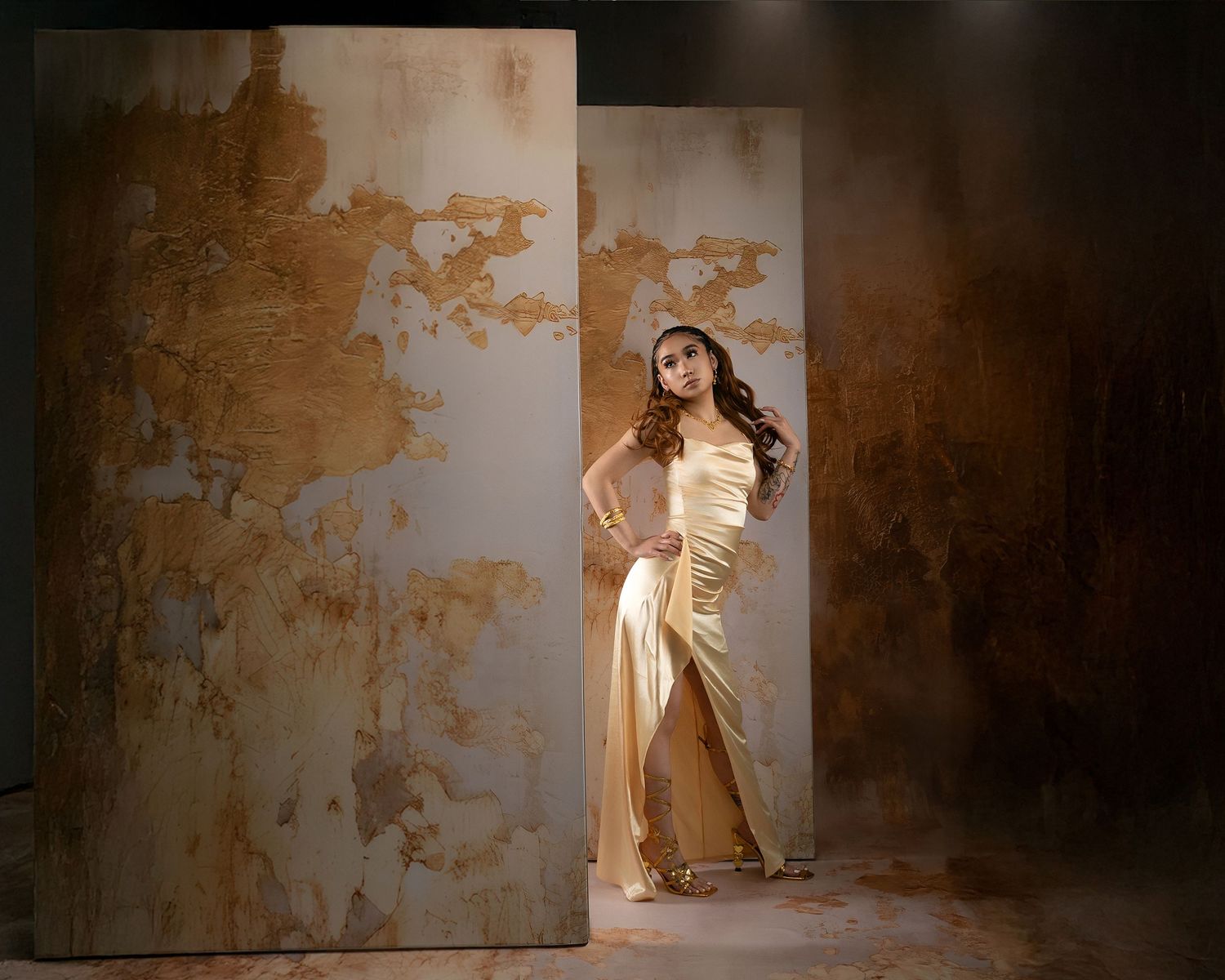 Elegant woman in gold satin gown poses dramatically between weathered textured wall panels in studio portrait.
