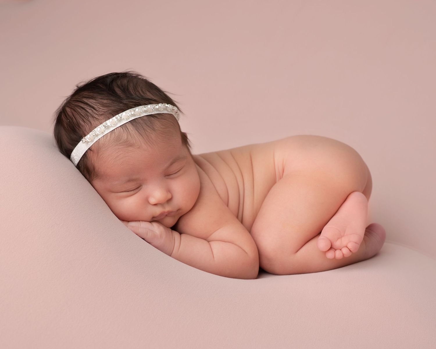Sleeping newborn baby girl with a delicate headband posed on a soft pink studio backdrop.