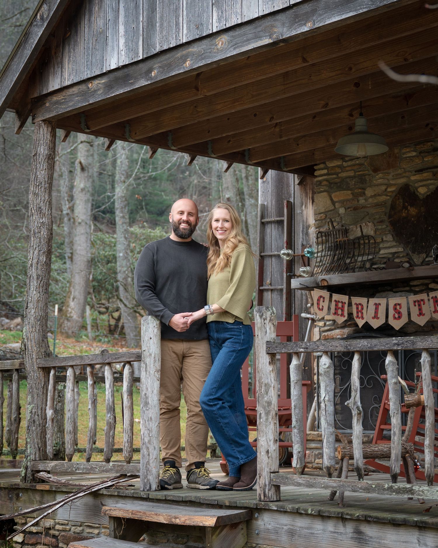 Couple smiling together on rustic cabin porch with Christmas banner in wooded outdoor setting.