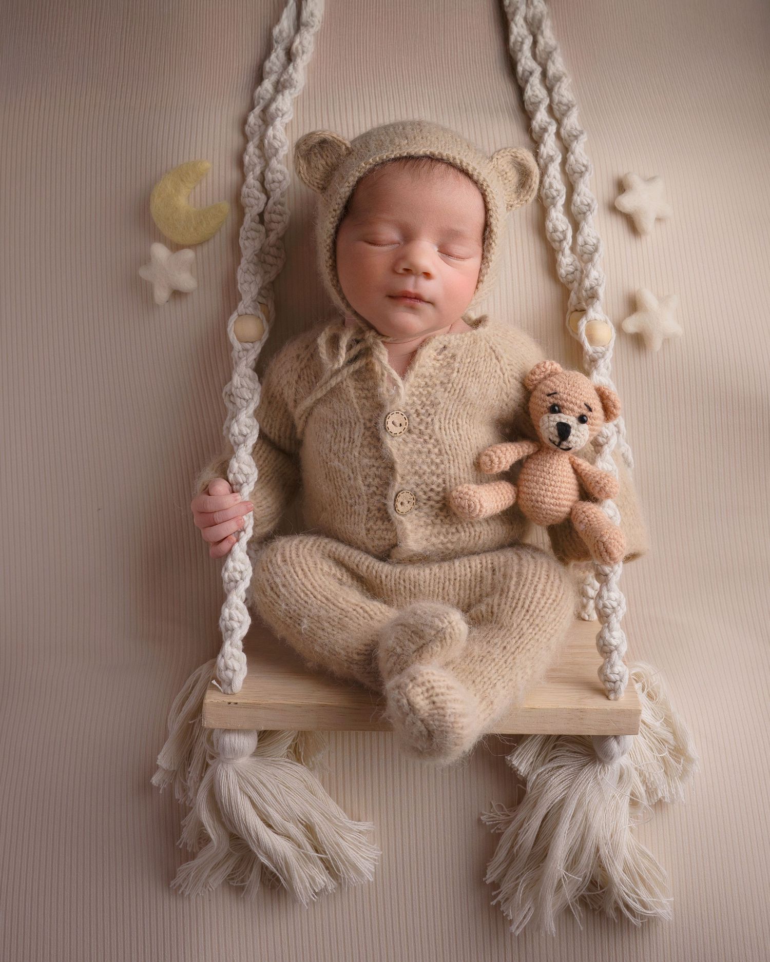 Newborn baby in knit bear outfit sleeping on a wooden swing with a teddy bear prop.