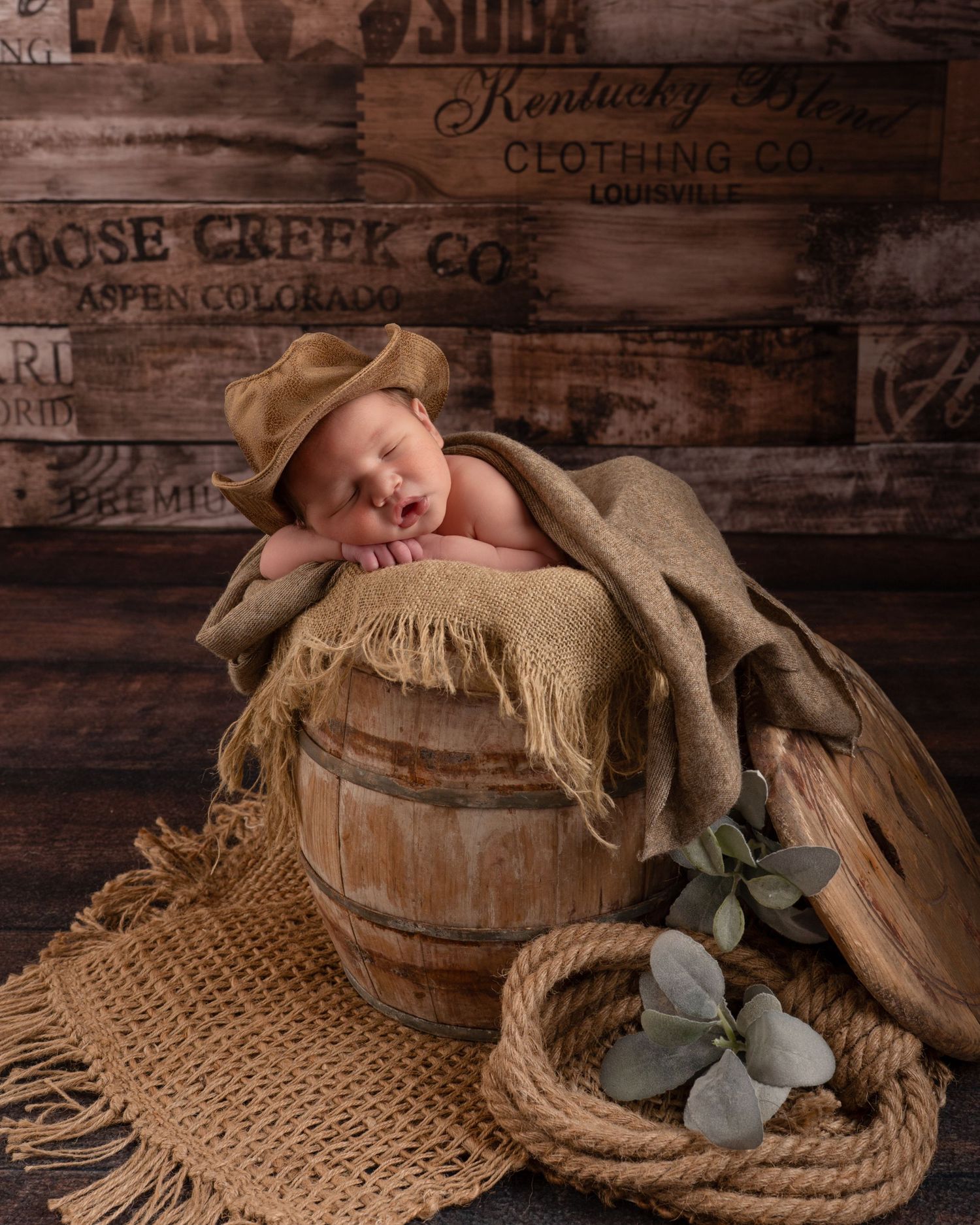 Newborn baby in a cowboy hat sleeping in a rustic wooden barrel with burlap on wood backdrop.