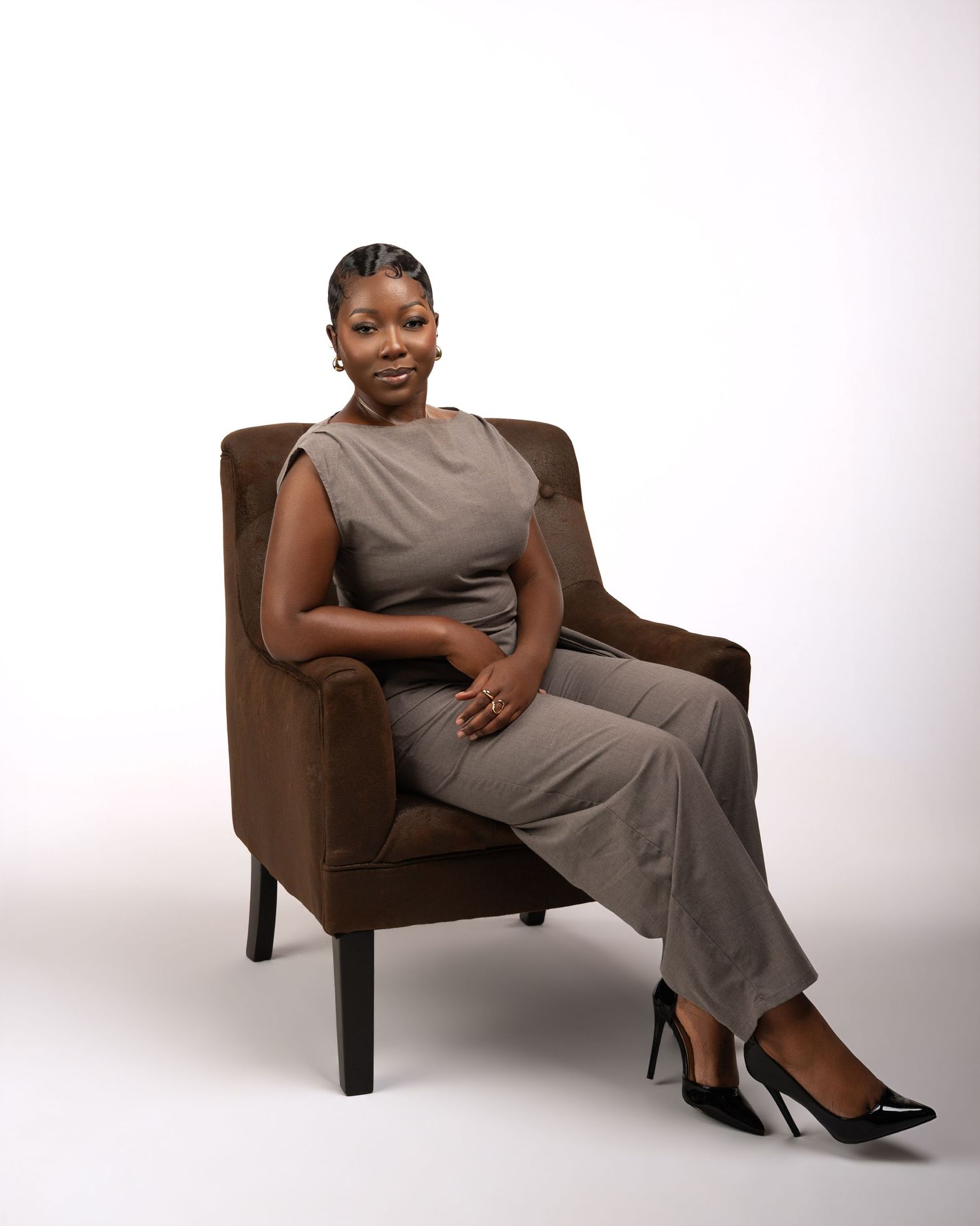 A confident Black woman in a gray jumpsuit and black heels sits poised in a brown velvet chair against a white background.